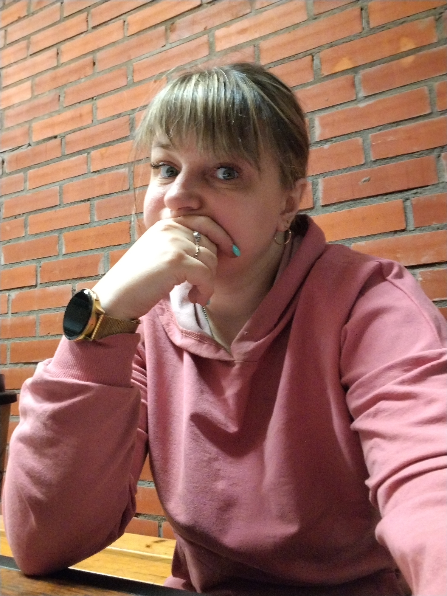 A medium close-up shot captures a woman sitting indoors, against a brick wall, in Шебунино, Russia. The woman, who appears to be an adult with light-colored hair and bangs, is looking directly at the camera with wide, light-colored eyes. Her left hand is raised to her mouth, partially covering it, giving her expression a sense of surprise, amusement, or shyness. She is wearing a pink hooded sweatshirt. On her left wrist, she has a gold-toned smartwatch with a mesh strap. A ring is visible on her ring finger, and her thumb sports teal nail polish. A small gold hoop earring can be seen in her right ear. The background consists of a reddish-brown brick wall with lighter mortar lines. In the lower part of the frame, a wooden surface, likely a table or bench, is visible where her arms rest. The lighting suggests an indoor setting with warm, artificial light. No visible text is present in the image.