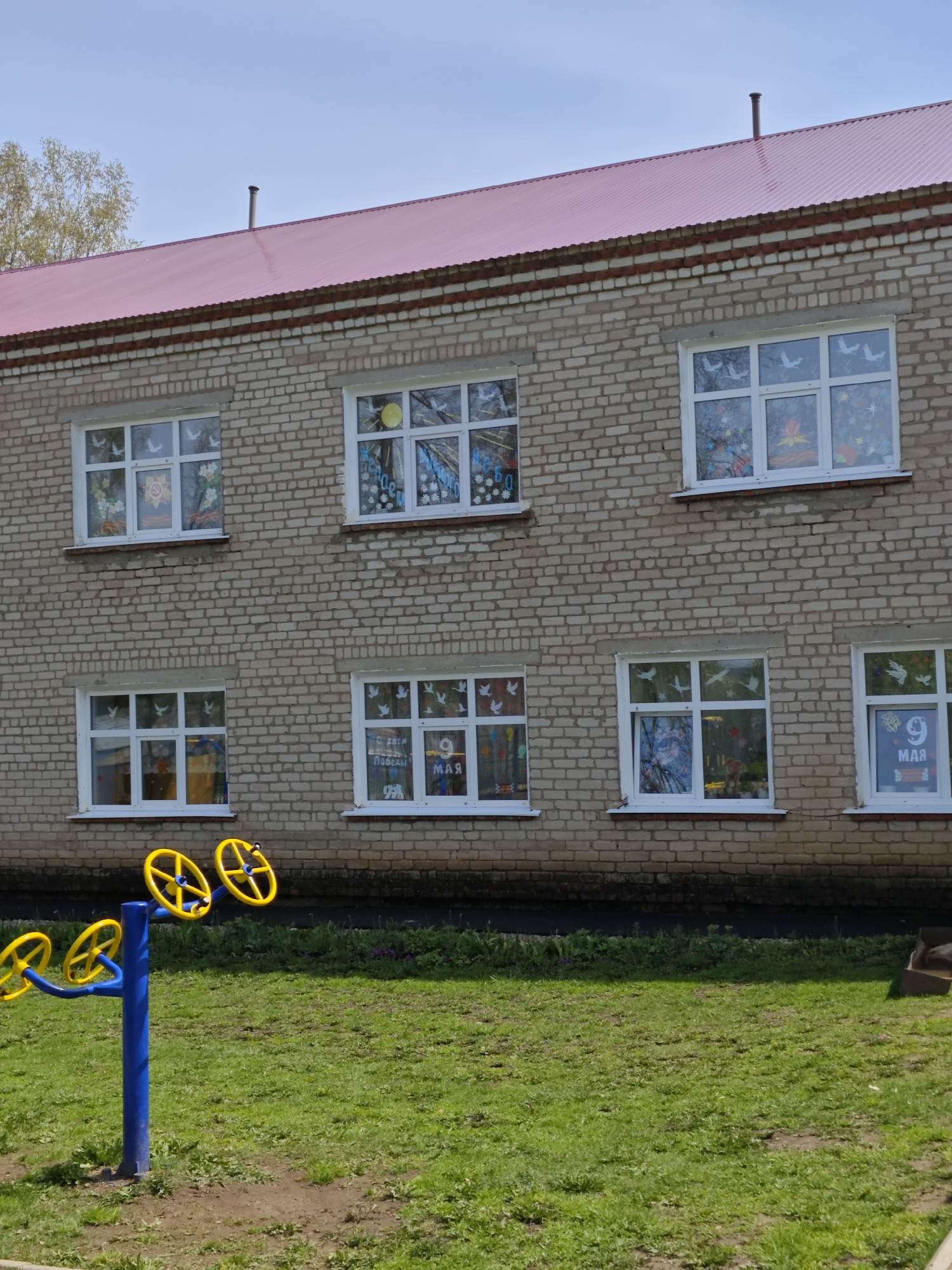 A two-story brick building with a red roof stands in Большая Уса, Russia. Six windows are visible, each decorated with various displays. The lower left window has a depiction of a yellow sun. Other windows show doves, flowers, and the date "9 МАЯ". In front of the building is a green lawn with playground equipment featuring blue supports and yellow wheels. The sky is partly cloudy.