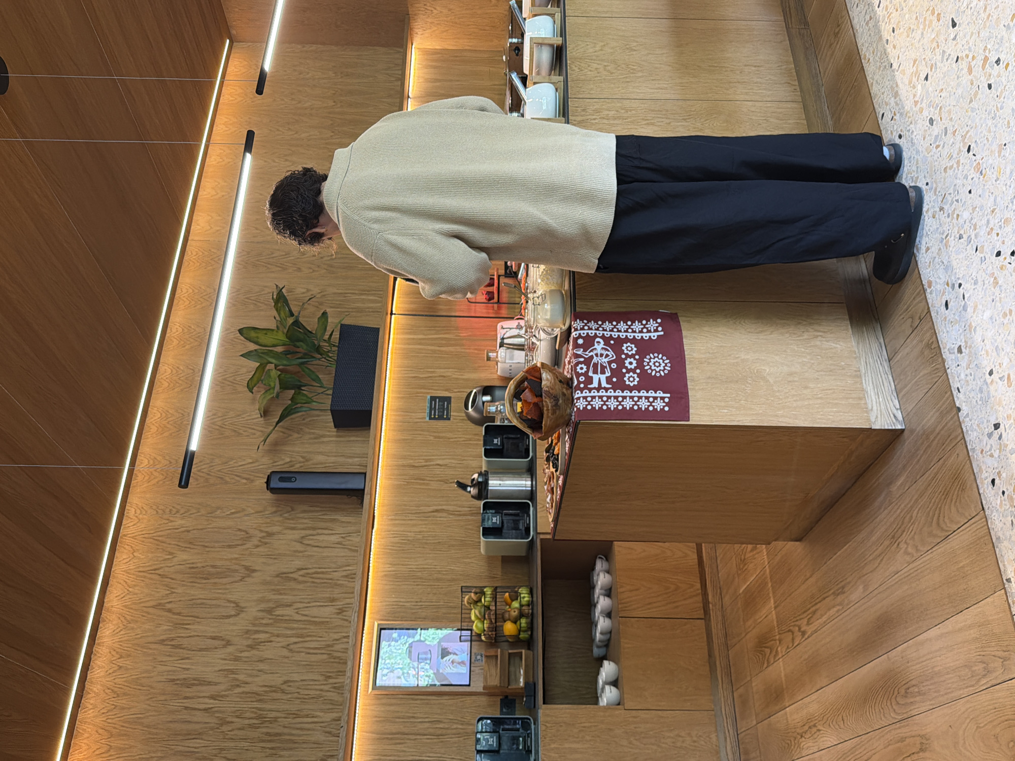 This is an indoor scene depicting a self-service beverage station or communal kitchen area. The dominant material in the room is light-colored wood paneling, which covers the walls and the base of the counter. The flooring is a mix of the same light wood planks and a light-colored, speckled terrazzo-like material.
In the center of the image, facing away from the viewer, stands a person with dark, curly hair. They are wearing a light beige or cream-colored ribbed sweater and loose-fitting black pants, paired with dark sandals or clogs. The person appears to be interacting with the items on the counter, possibly preparing a drink or snack.
The counter features several appliances, including what appear to be coffee machines or kettles, and various mugs or cups arranged on wooden shelves. A small, dark woven basket filled with dried fruit or nuts sits next to one of the machines. Below the main counter, on a lower shelf, there is a wire basket brimming with fresh fruit, including green apples and oranges. A small digital screen on the wall displays a vibrant image, possibly of nature or an advertisement.
A striking red placemat with a white, intricate decorative pattern and a stylized human figure is laid out on the counter to the right. The walls are illuminated by modern, linear LED light strips; some are embedded horizontally in the wood panels, casting a warm glow, while others hang vertically from the ceiling. A dark, rectangular speaker-like device is mounted on the wooden wall to the left of the counter. The overall atmosphere suggests a contemporary, well-lit, and functional space, possibly in an office, hotel, or co-working environment. No discernible text is clearly visible in the image.