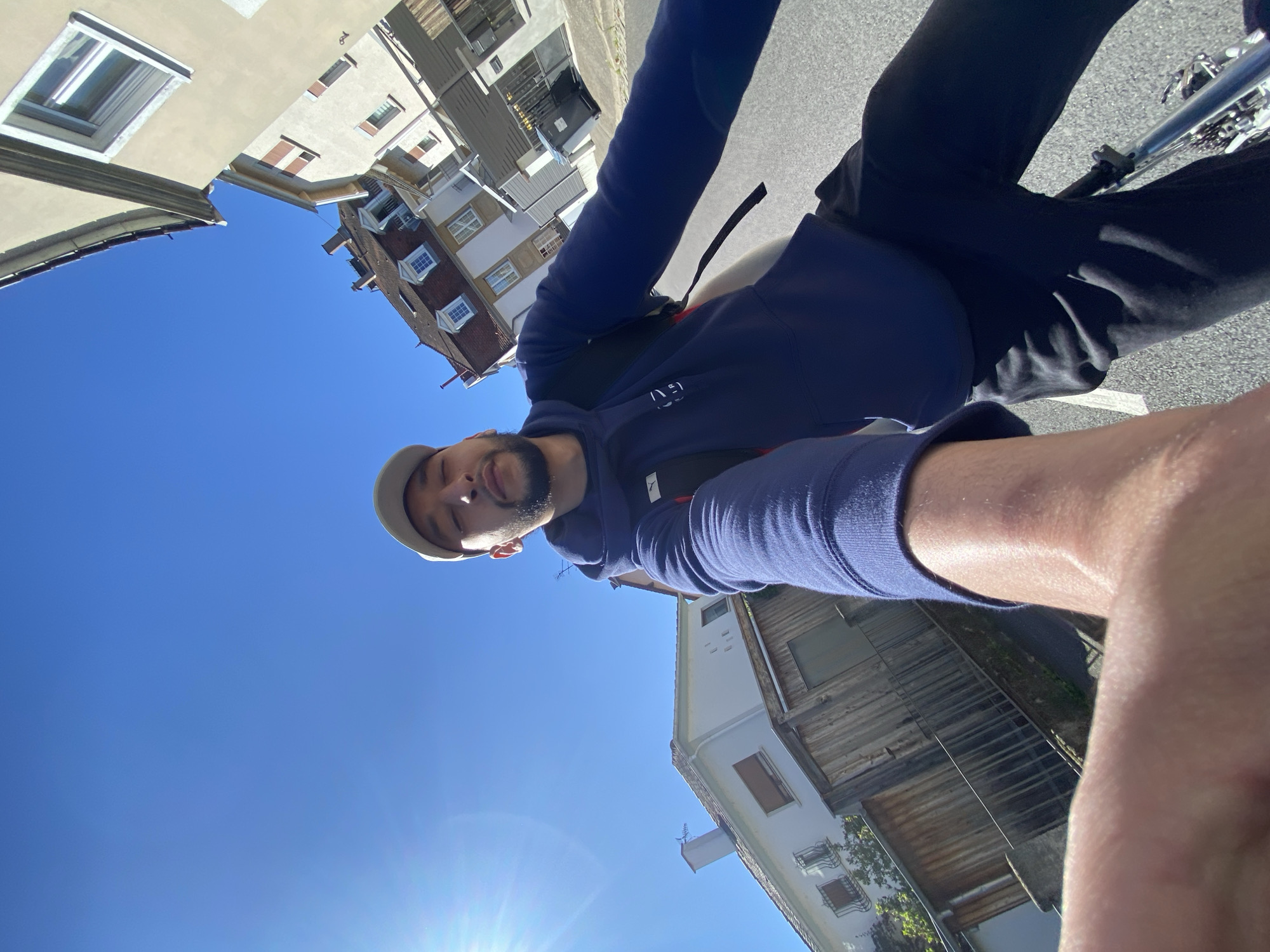 A person is on a bicycle on a street in Hoenheim, France. The person wears a blue hoodie, dark pants, and a light-colored baseball cap. Buildings and a clear blue sky are visible in the background.