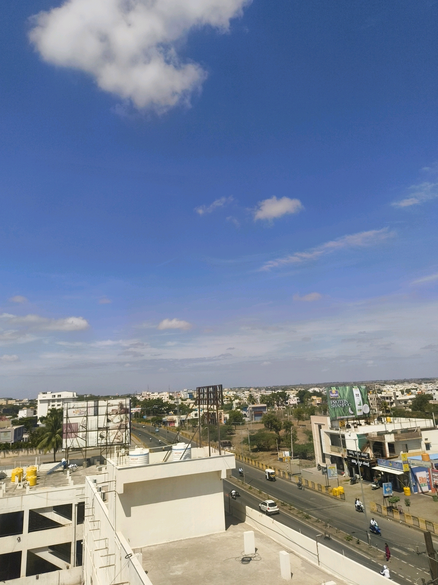 The image captures a wide-angle view of a cityscape in Vijayapura, India, under a bright blue sky dotted with scattered white clouds. In the foreground, the rooftops of modern buildings are visible, some featuring water tanks and solar panels. A road runs through the middle ground, carrying light traffic including cars, motorcycles, and an auto-rickshaw. Buildings line the road, with shops and advertisements visible. The background reveals a dense urban landscape with numerous houses and trees extending to the horizon. A large billboard advertising "PROTEIN SHAMPOO" is prominent on one of the buildings. Several individuals are present on the sidewalk and on the road, engaged in daily activities. The overall scene depicts a typical day in an Indian city.
