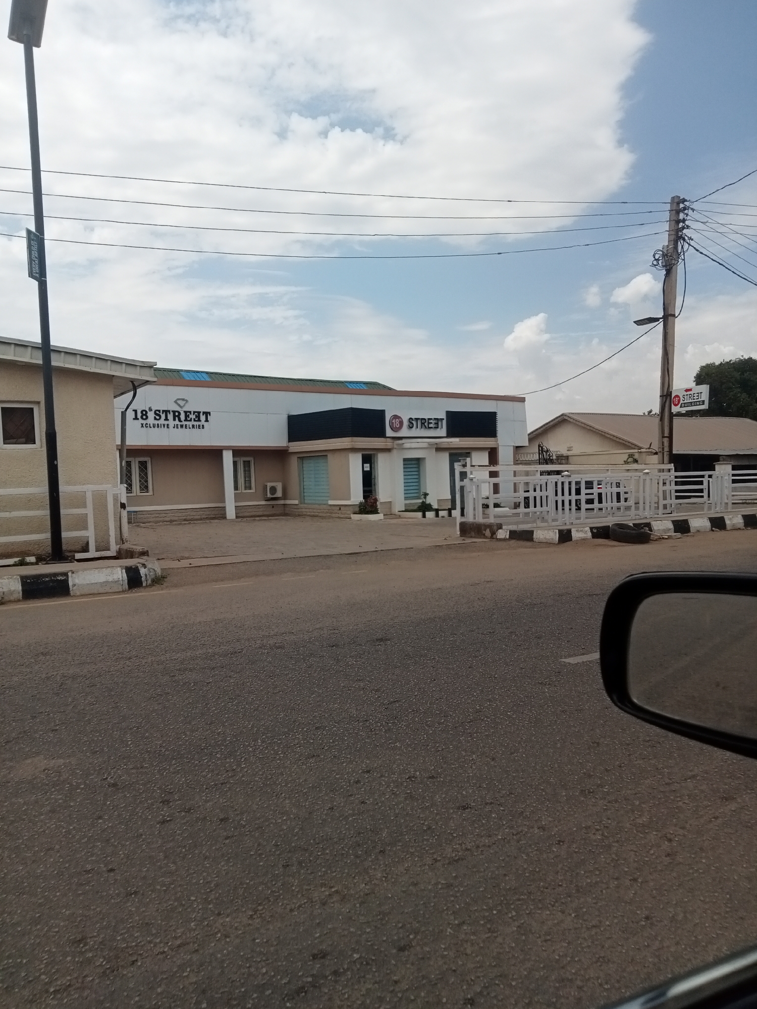 The image shows a building in Jos, Nigeria identified as "18th Street Exclusive Jewelries". The building is a one-story structure with a light beige and white facade. There are signs displaying the "18th Street" name on the building itself and on a signpost near a utility pole. A white fence surrounds part of the property. The foreground shows a paved road and a car's side mirror is visible in the bottom right corner. The sky is partly cloudy.