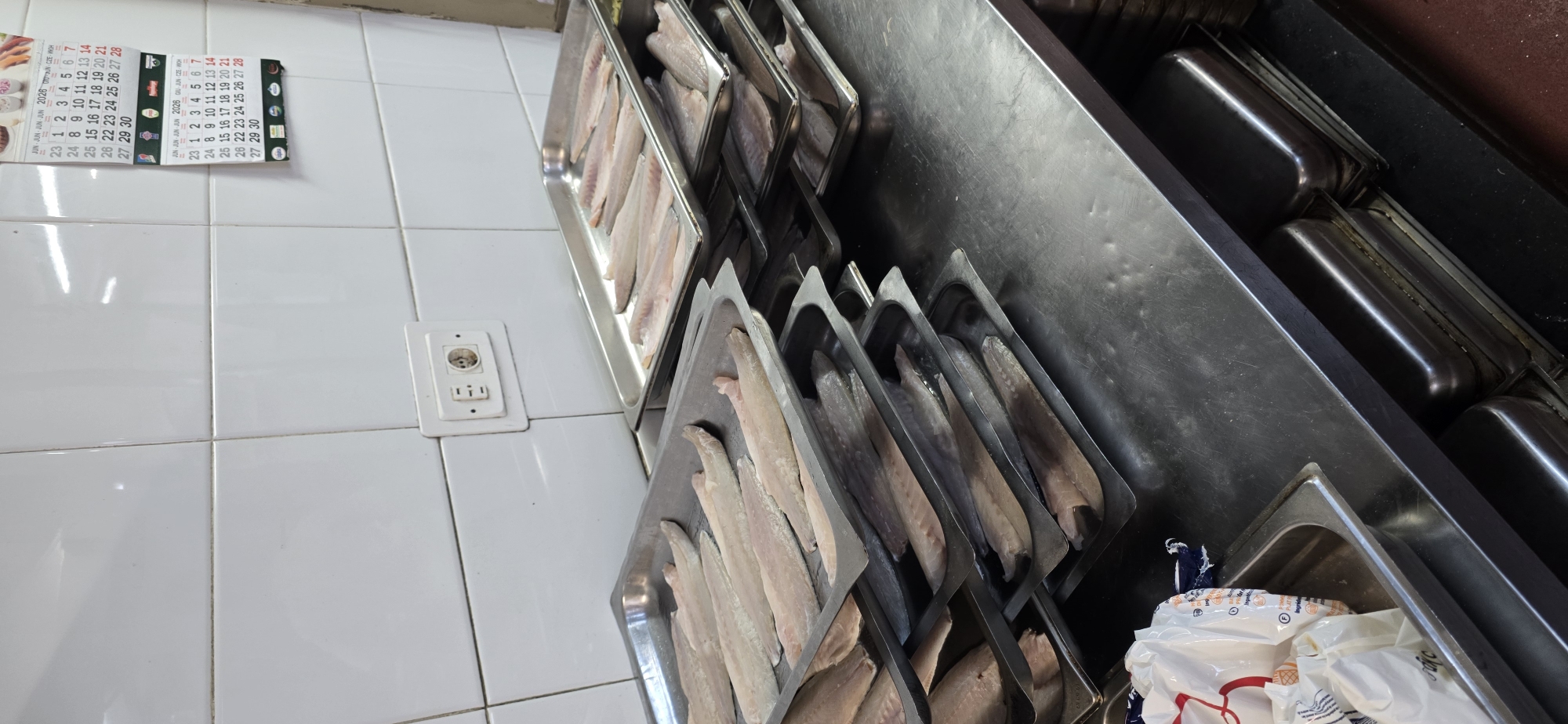 In a commercial kitchen environment in Sallent de Gállego, Spain, multiple stainless steel trays are stacked and filled with raw fish fillets. The fish appears to be white fish, such as cod or hake, and is arranged in single layers within the trays. The kitchen has white tiled walls and a stainless steel countertop, a common setup for food preparation areas. An electrical outlet is visible on the wall, as is a calendar hanging above the trays, indicating the current date is within the month of June in the year 2026. The overall impression is of a food preparation area ready for cooking or further processing of the fish.
