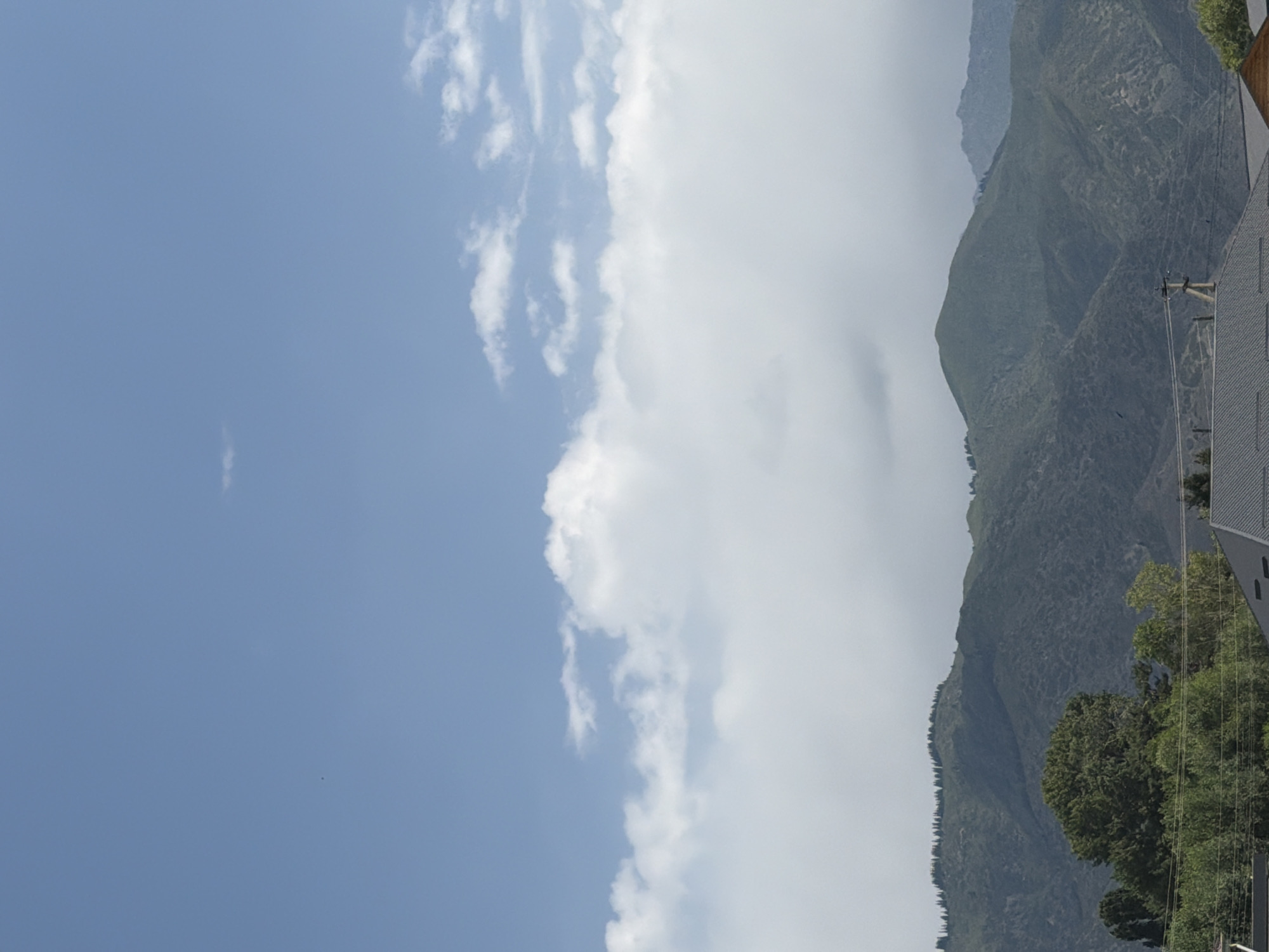 The image captures a vast, clear blue sky with scattered white clouds, dominating the upper portion of the frame. Below the sky, a band of dense, white clouds stretches horizontally, partially obscuring the mountain range in the background. These mountains are rugged and covered in dark green vegetation, with a distinctly textured surface suggesting varied terrain. The lower part of the image shows a line of trees in the foreground, with a large, dense tree on the left and a cluster of smaller trees to its right. In the very bottom of the image, the roofs of several buildings are visible, one with a dark gray, gabled roof and another with a lighter brown, angular roof. The scene appears to be a landscape, likely on the outskirts of a city, given the presence of buildings and utility poles with wires.