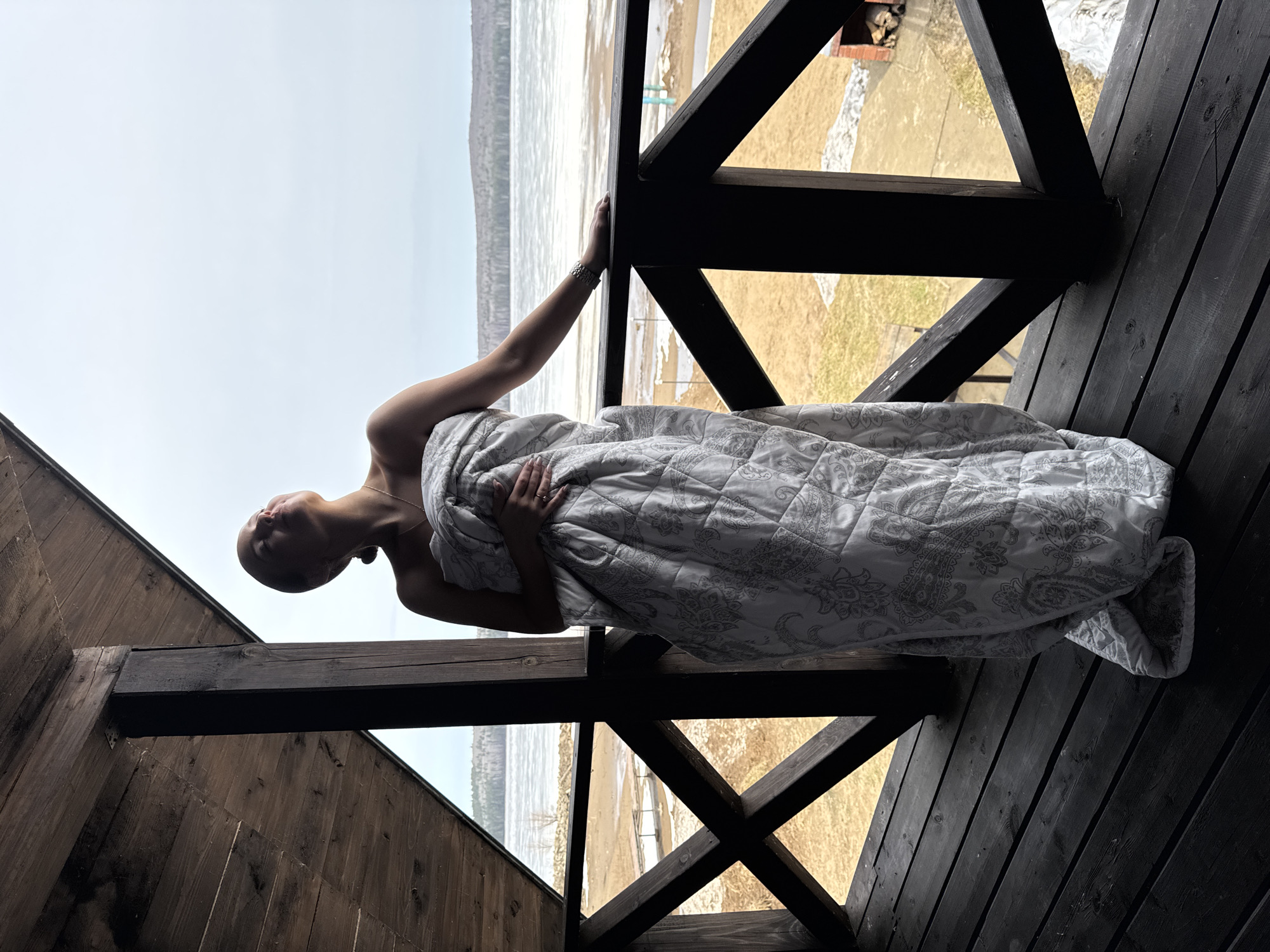 A bald woman is standing on a dark wooden balcony, wrapped in a white patterned blanket. She is leaning against the railing, looking up. In the background, through the wooden beams of the balcony, there's a view of a lake with a sandy beach and a partially frozen surface. The sky is overcast. Based on visual evidence, this scene appears to be located in Chaikovsky, Russia.