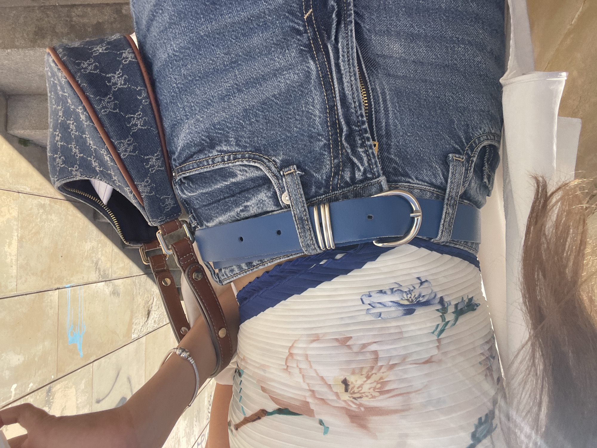 The image shows a person's torso, wearing blue jeans with a blue belt over a white, floral-patterned dress. The person is carrying a patterned denim handbag with brown leather straps. The image also includes a partial view of the person's arm with a silver bracelet. The background appears to be a stone surface, possibly stairs, with some graffiti. This is probably taken in the city of València, Spain.