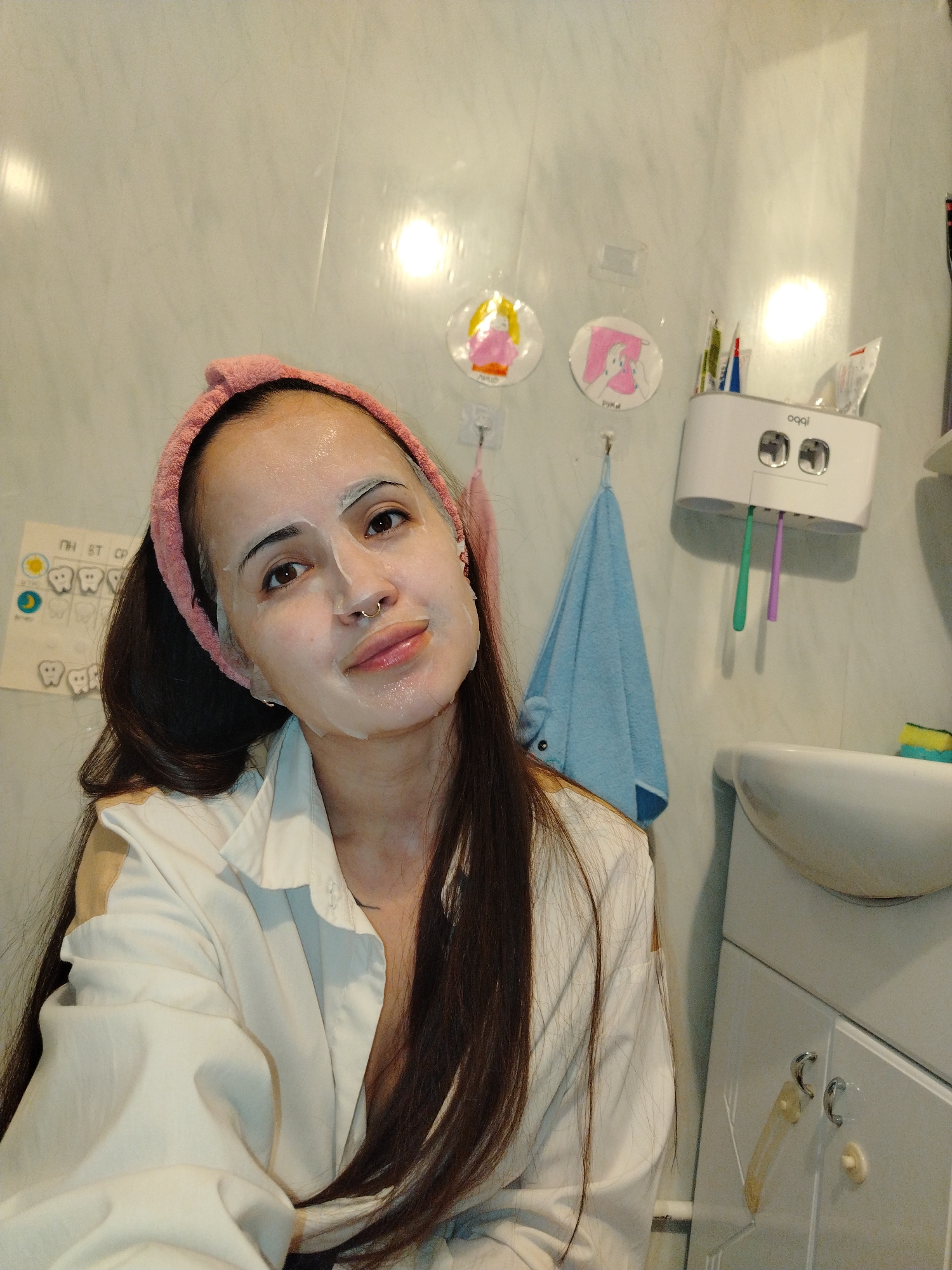 The image shows a woman in a bathroom. She is wearing a pink headband and a white sheet face mask. She is also wearing a white shirt. The bathroom has white tile walls and a white vanity with a sink. There is a toothbrush holder and a blue towel on the wall. There are also some drawings on the wall.