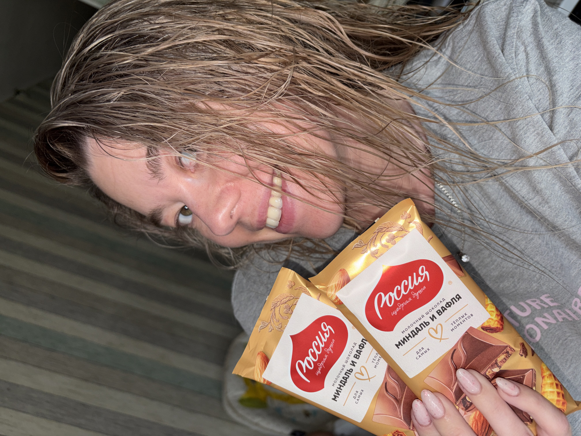 A woman with wet, blonde hair and a wide smile is holding two bars of "Russia" milk chocolate with almonds and waffles. She is wearing a gray sweatshirt. The backdrop consists of a wall with wooden panels and a glimpse of a "CONAIR" branded object in the background, which might indicate a domestic setting. Given the chocolate packaging, it is plausible that the photo was taken in a location where the product is sold or consumed, potentially in Russia.