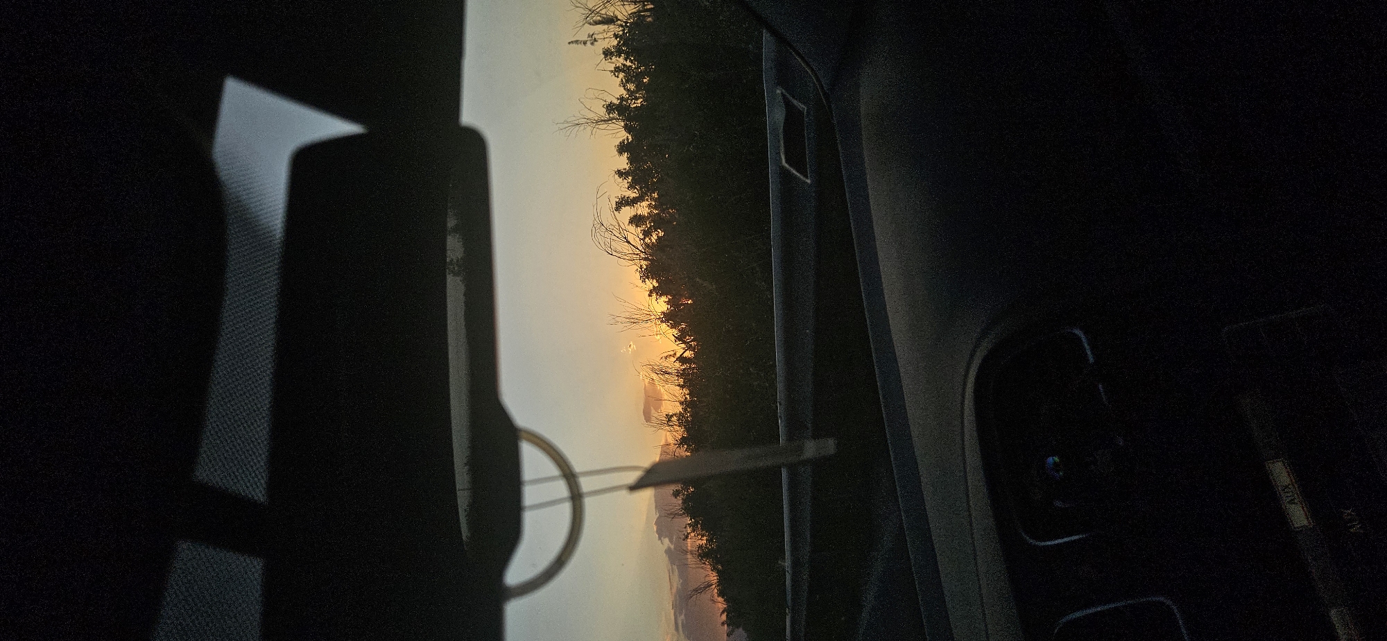 The image shows a view from inside a vehicle, likely a car, at sunset. Outside, there are trees and a glimpse of mountains visible in the distance. The sun is setting, casting a warm glow on the scene. The interior of the car is mostly dark, with parts of the dashboard and rearview mirror visible in silhouette. There's no indication of Kovachevo, Bulgaria. No people are visible.