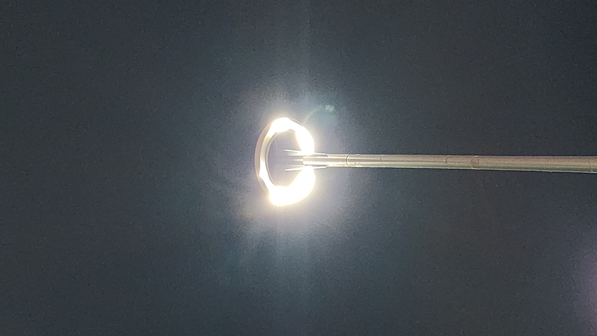 A streetlamp with a circular, illuminated head stands against a dark, possibly night sky. The lamp is positioned in the center of the frame, extending from the bottom to the top. The sky appears to be a uniform dark gray, with subtle variations in tone. The streetlamp itself is metallic and has a segmented pole. The head of the lamp emits a bright, white light that creates a halo effect around it, with some lens flare visible. There are no people or other objects clearly visible in the frame. The overall impression is of a solitary, illuminated object in a dark environment. The image was taken in the city of Yerevan, Armenia.