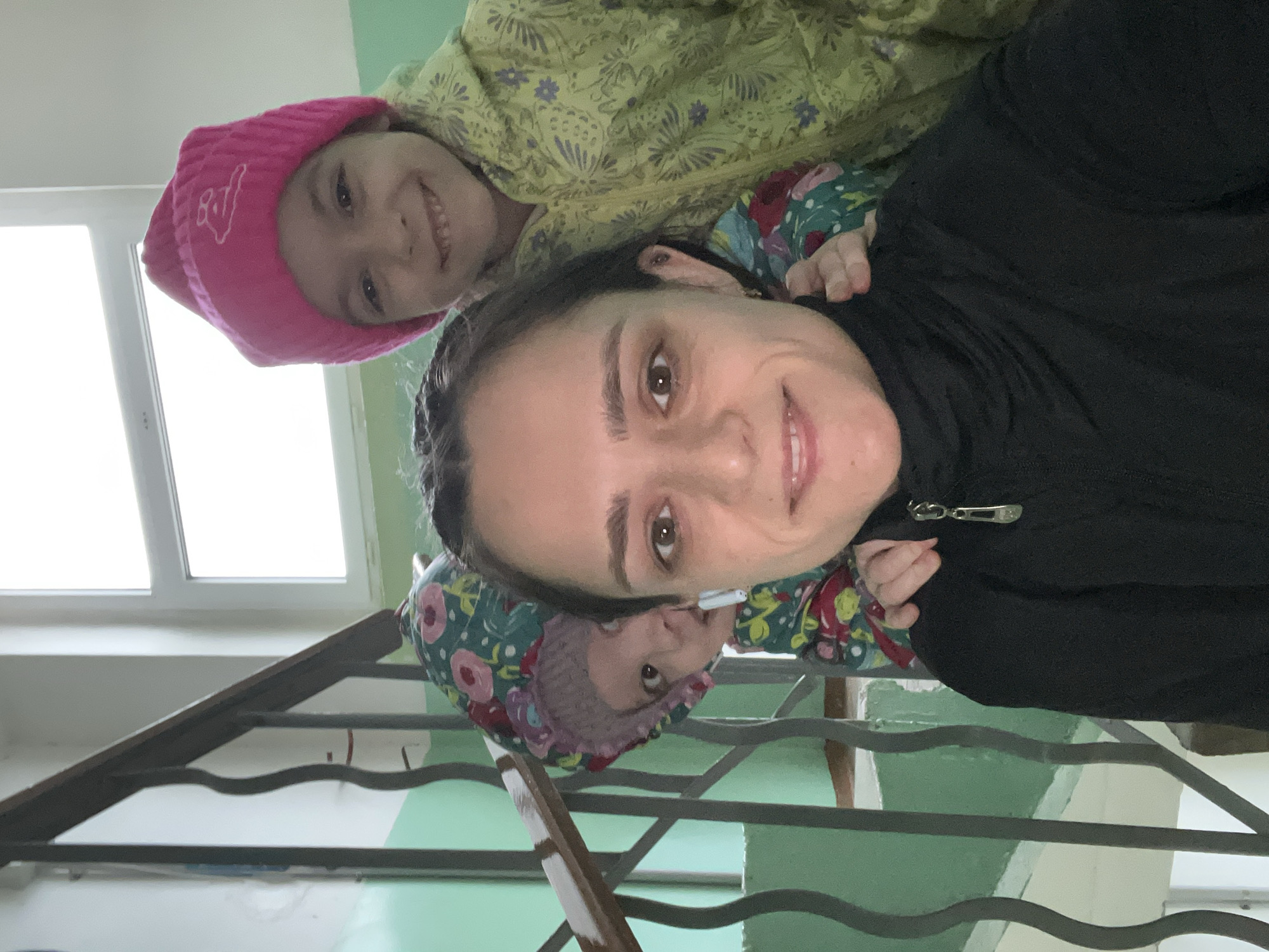 A medium-shot photo shows a woman, a young girl in a pink hat, and a small child on a staircase in what appears to be an apartment building. The woman is looking at the camera and smiling. The young girl is smiling as well. The smaller child is peeking out from behind the woman's shoulder. The woman is wearing a black jacket. The young girl is wearing a yellow patterned jacket. The smaller child is wearing a floral patterned jacket and hat. The staircase has metal railings and light green walls. A window is visible in the background.
I am unable to ascertain the location, Chaikovsky, Russia, from this image.