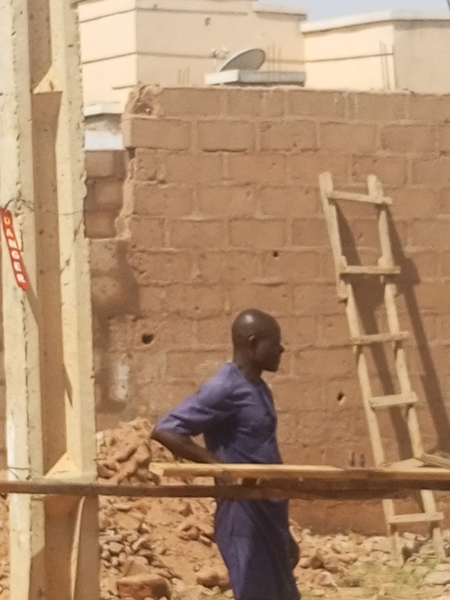 The image depicts an outdoor scene at what appears to be a construction or renovation site in Tempure, Nigeria, under bright daylight.
The primary subject is a man, shown in profile, facing towards the right side of the frame. He has a bald head and is dressed in a long-sleeved, loose-fitting blue or purple tunic. His right arm is bent, with his hand resting on a robust horizontal wooden plank or beam that runs across the foreground, creating a barrier between the viewer and the man. His posture suggests he is either resting, observing the work, or taking a break.
Behind the man is a reddish-brown brick wall, which appears to be unplastered or in a state of partial disrepair. Leaning against this wall on the right side of the image is a simple, rustic wooden ladder. The ground beneath is uneven and covered with loose dirt, small rocks, and construction debris.
On the far left, several vertical concrete or wooden pillars are visible, possibly part of scaffolding or a building's framework. A red tag, clearly bearing the word "DANGER" written vertically in white letters, is attached to one of these pillars.
In the background, above the brick wall, parts of light-colored, possibly beige or cream-colored, buildings with flat roofs can be seen under a clear, bright sky. One of these buildings features a satellite dish on its roof. The strong sunlight casts prominent shadows, particularly from the ladder onto the brick wall, indicating that the time of day is likely midday or early afternoon.