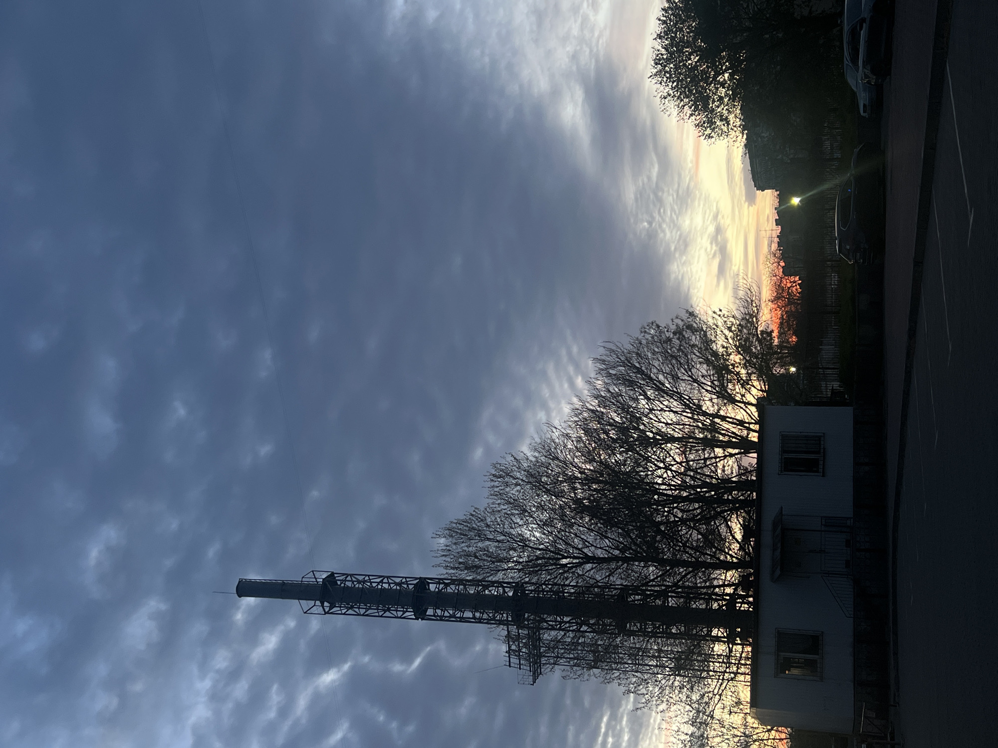 The image shows a vertical view of a landscape under a cloudy sky. On the right, a building is partially visible, standing next to trees with bare branches. To the left of the building, there is a tall metallic tower, likely a chimney or observation tower, extending upwards into the sky. The sky is filled with clouds, and a glow of orange or yellow is visible in the distance, possibly indicating sunrise or sunset. In the upper right corner, other trees, possibly part of a park, are visible, along with silhouettes of cars parked nearby. The scene appears to be taken at dusk or dawn. Based on the presence of the tower and industrial structures, it is possible this image was taken in the city of Odesa, Ukraine, due to Odesa's industrial facilities.