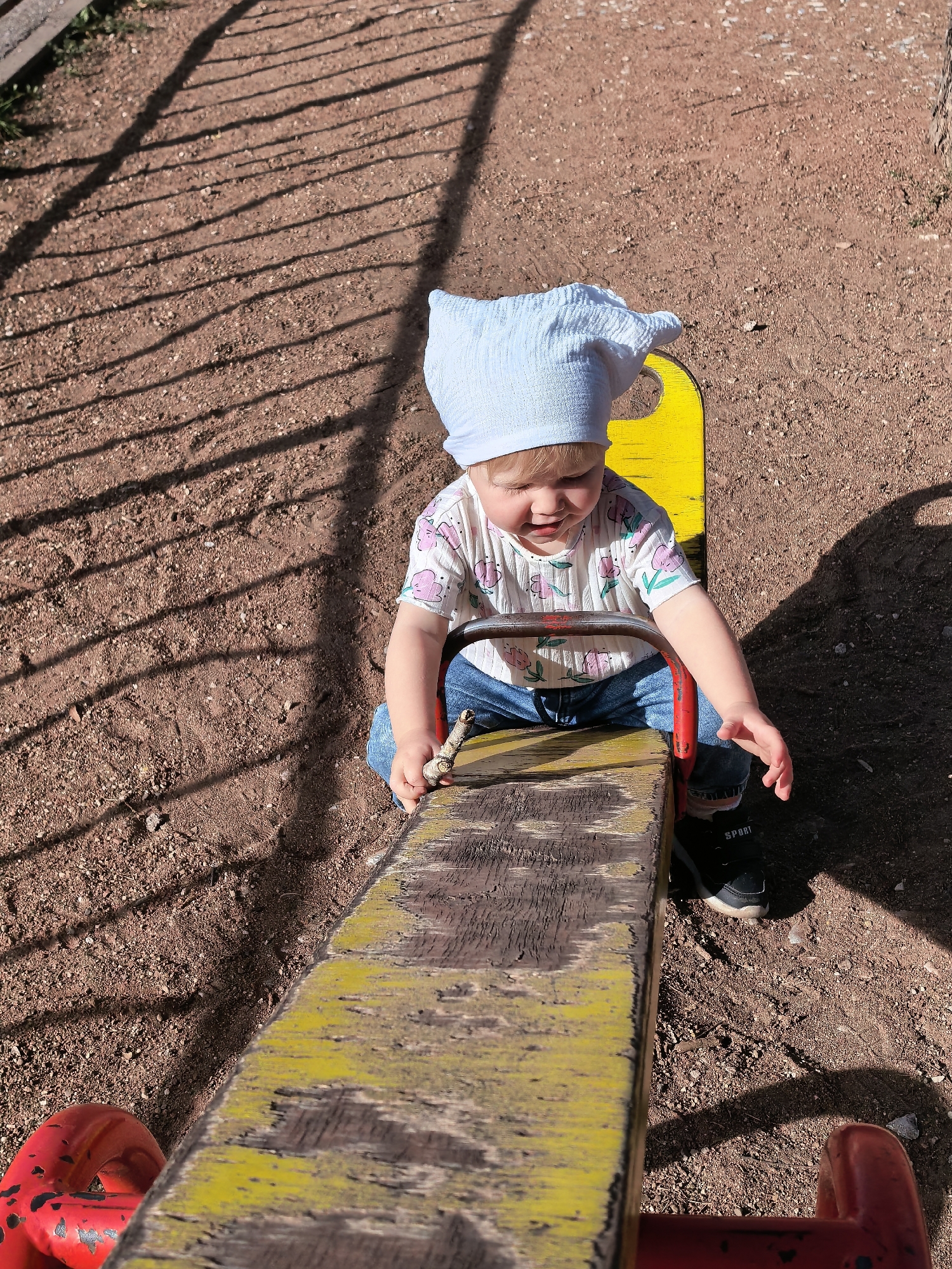 A young child, wearing a light blue beanie, a white t-shirt with pink floral patterns, and blue jeans, sits on a weathered yellow and red seesaw. The child is holding a small stick in their right hand. The seesaw is on a ground covered with reddish-brown dirt and small pebbles. Sunlight casts striped shadows from an unseen fence onto the ground. The scene is outdoors, likely in a park or playground, with no specific landmarks indicating the city of Sterlitamak, Russia.