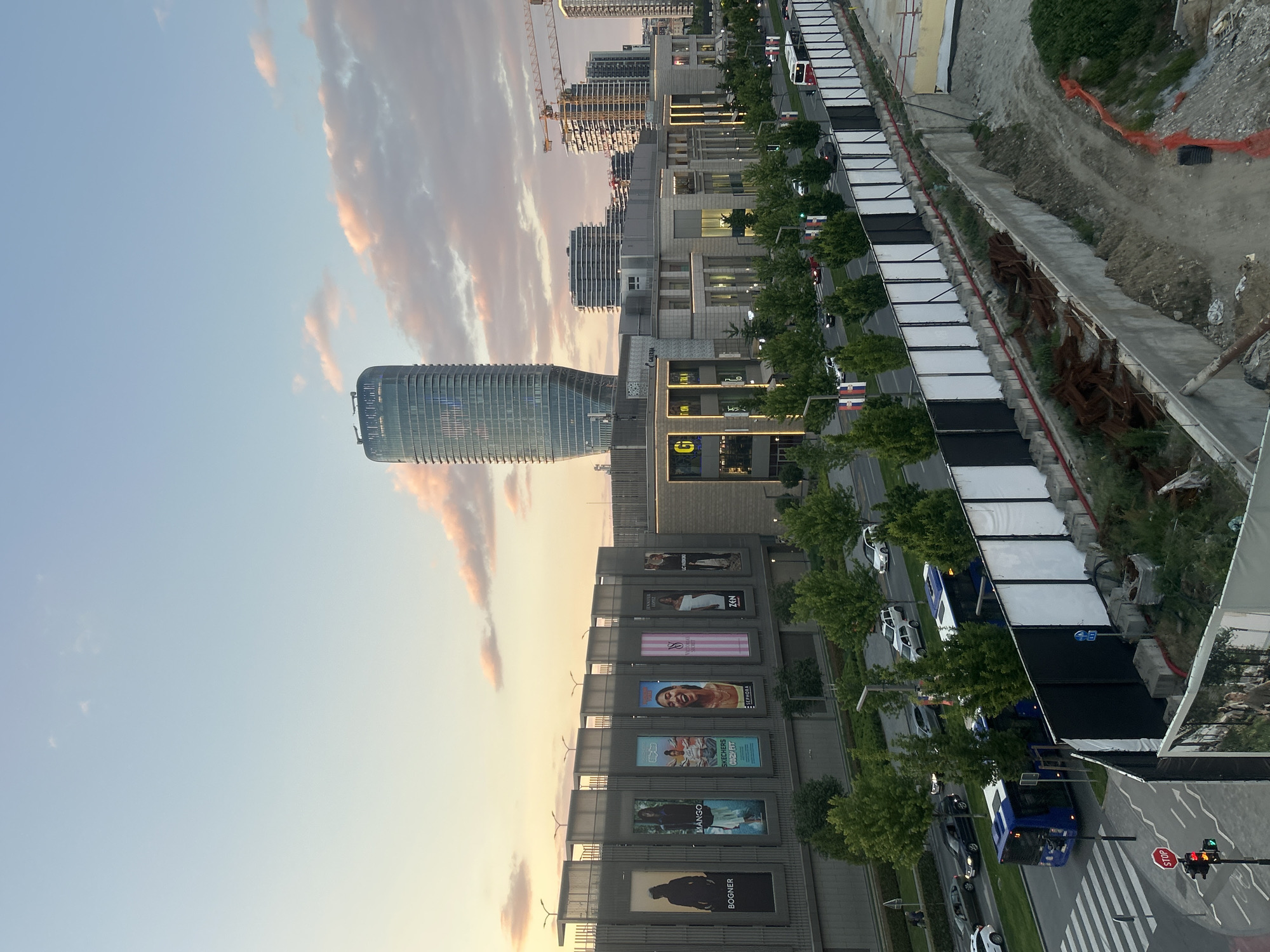 The image shows a view of Belgrade Waterfront in Belgrade, Serbia. A modern, cylindrical skyscraper dominates the left side of the frame, set against a backdrop of a sky with pastel-colored clouds. To the right of the skyscraper is a series of modern buildings with retail spaces and large billboards advertising various brands.
Trees line the pedestrian walkways and roads, adding greenery to the urban landscape. A construction site is visible on the right side, with exposed earth and building materials. In the lower right corner, there is a street with vehicles, including a bus, and a traffic light signaling green. A stop sign is also visible.
The overall impression is of a contemporary urban development.