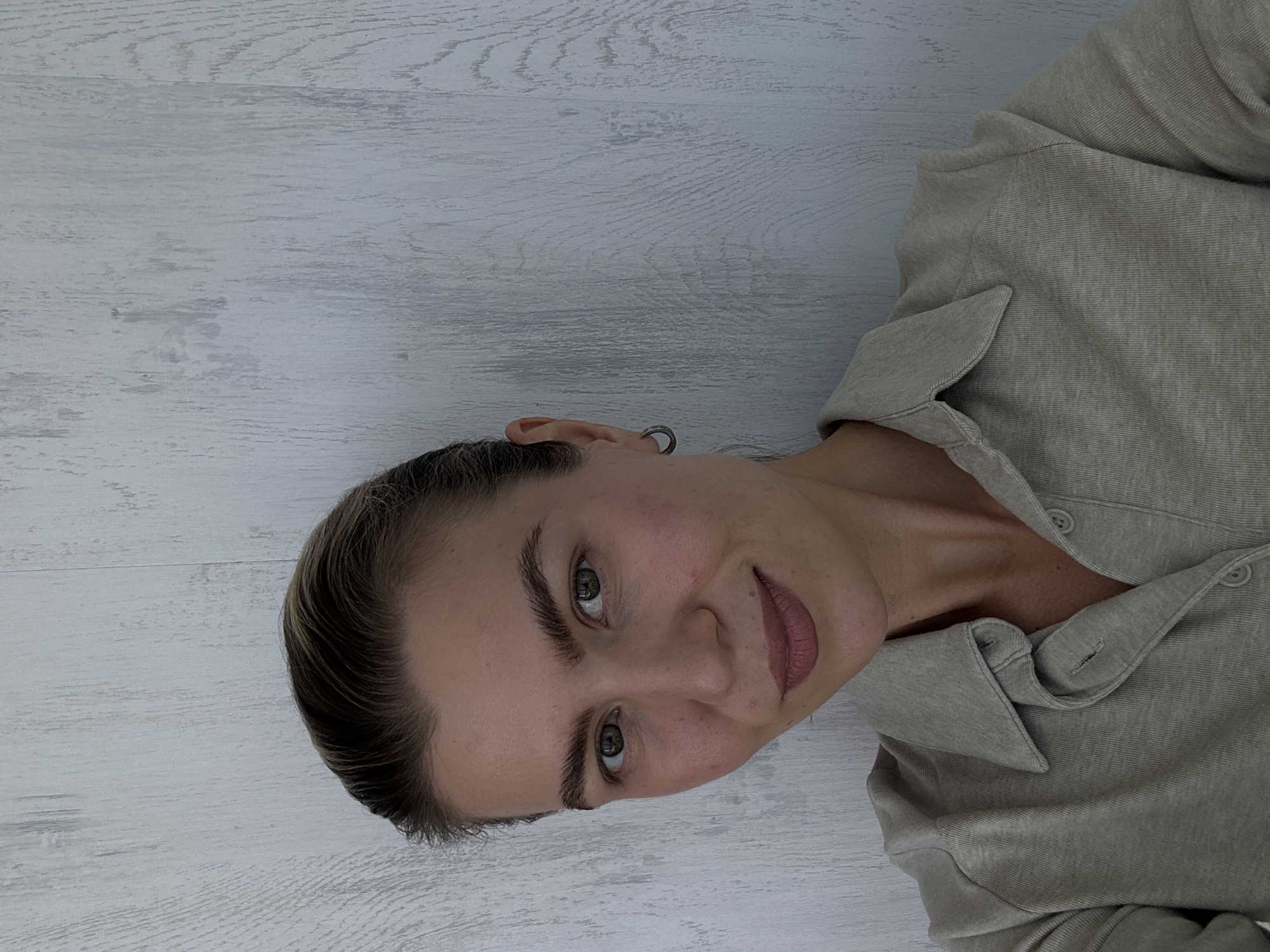 The image shows a woman lying on her left side, looking directly at the camera with a gentle, slight smile. Her head is positioned towards the lower left of the frame, and her body extends towards the upper right. She has dark hair pulled back from her face, revealing her ears. A small, silver-toned hoop or cuff earring is visible on her right ear (viewer's left). Her eyes are hazel or greenish, and her eyebrows are dark and well-defined. Her lips are adorned with light pink or mauve lipstick and are slightly parted. She has a natural, clear complexion. She is wearing a long-sleeved, collared shirt in a light beige or khaki color, made of a subtly textured fabric, possibly linen or cotton, with visible buttons down the front. The background is a light-colored, possibly whitewashed or bleached wooden surface, showing distinct grain patterns, suggesting an indoor setting like a floor or a wall. The lighting is soft and even, highlighting her features. No text is visible.