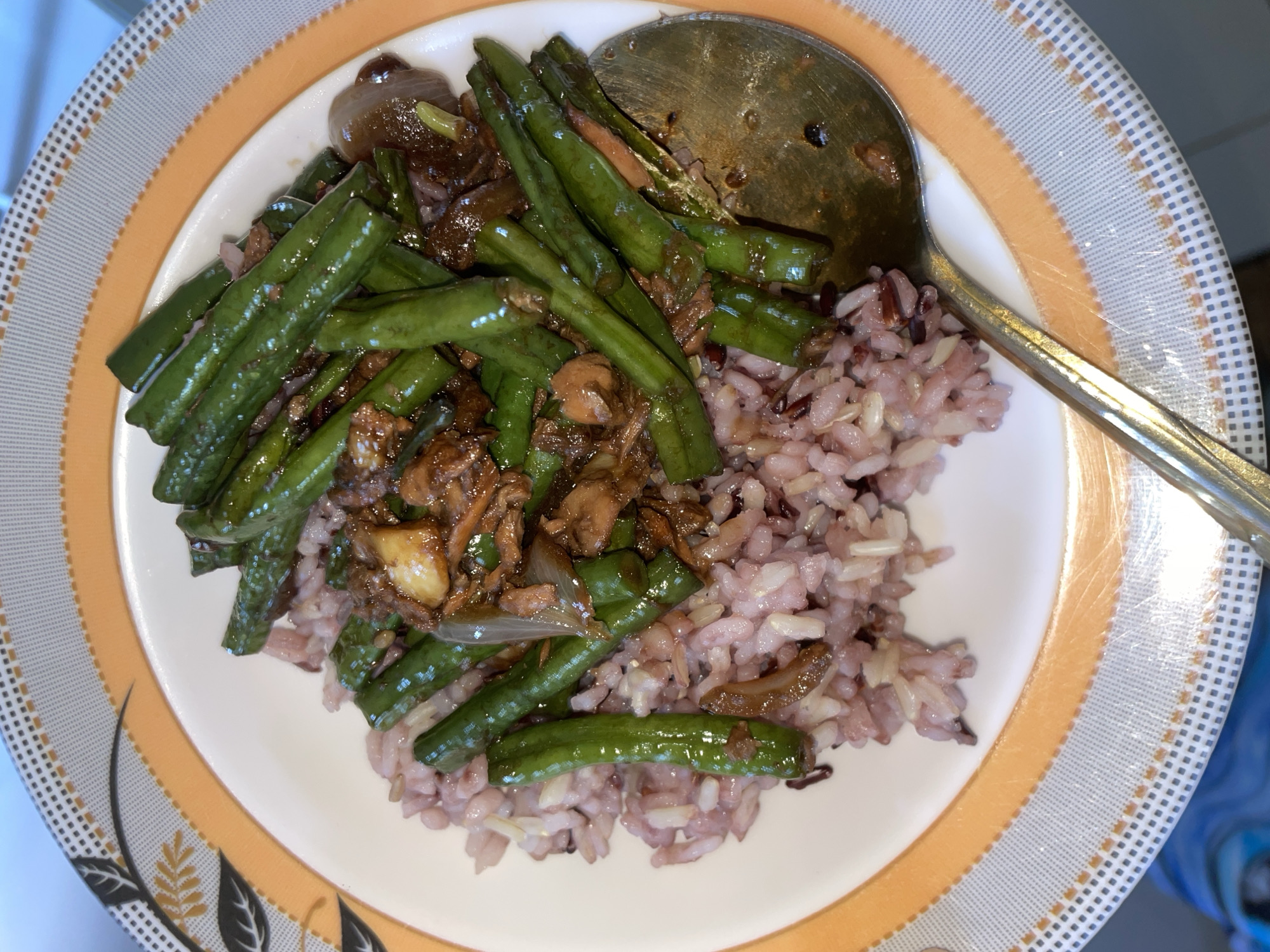 A plate of food is shown, featuring a portion of purple and white rice topped with stir-fried green beans and what appears to be shredded meat, possibly beef or chicken, in a dark sauce. A metallic spoon rests on the right side of the plate, partially in the food. The plate has an orange and grey patterned rim. The background is blurred, suggesting an indoor setting. There are no discernible landmarks indicating Davao City, Philippines.
