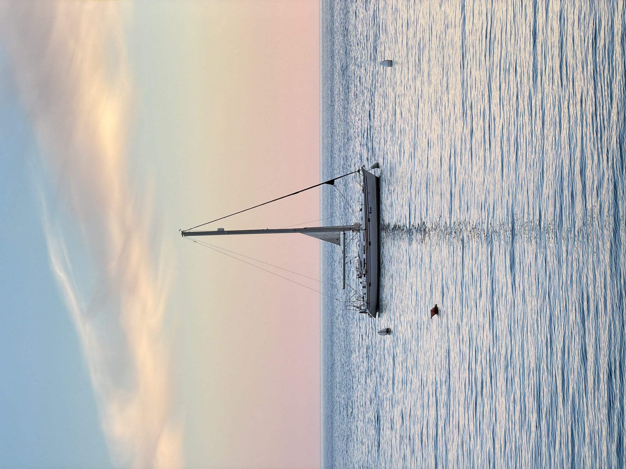 The image depicts a sailboat at anchor in calm water, with the sky transitioning from blue to pastel shades of pink and yellow in the background. The sailboat is white with a tall mast and rigging. The water has gentle ripples, with a buoy visible near the boat. The red flag is also near the boat. The location is identified as Petrovac centar, Montenegro.