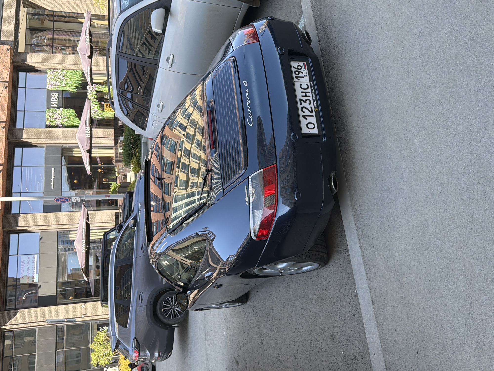 A close-up, slightly rotated, outdoor shot features the rear of a dark blue Porsche Carrera 4 parked on a street. The car is parked adjacent to a light gray SUV and a white vehicle. The license plate of the Porsche is clearly visible and reads "O123HC 196". The reflections of surrounding buildings are prominent on the car's surface.
In the background, a building with large windows and brick accents is visible. There is a sign that reads "АРЕНДА площадь 102 м² 8 (958) 224 30 41" and restaurant signs that read "ИВА". Additionally, patio umbrellas and greenery are visible in front of the building.
Based on the license plate, and the Cyrillic text on the building signage and the license plate itself, the location is likely in Russia. The 196 region code suggests that the location is in or near Moscow or St. Petersburg, however, other regions utilize the 196 code as well.