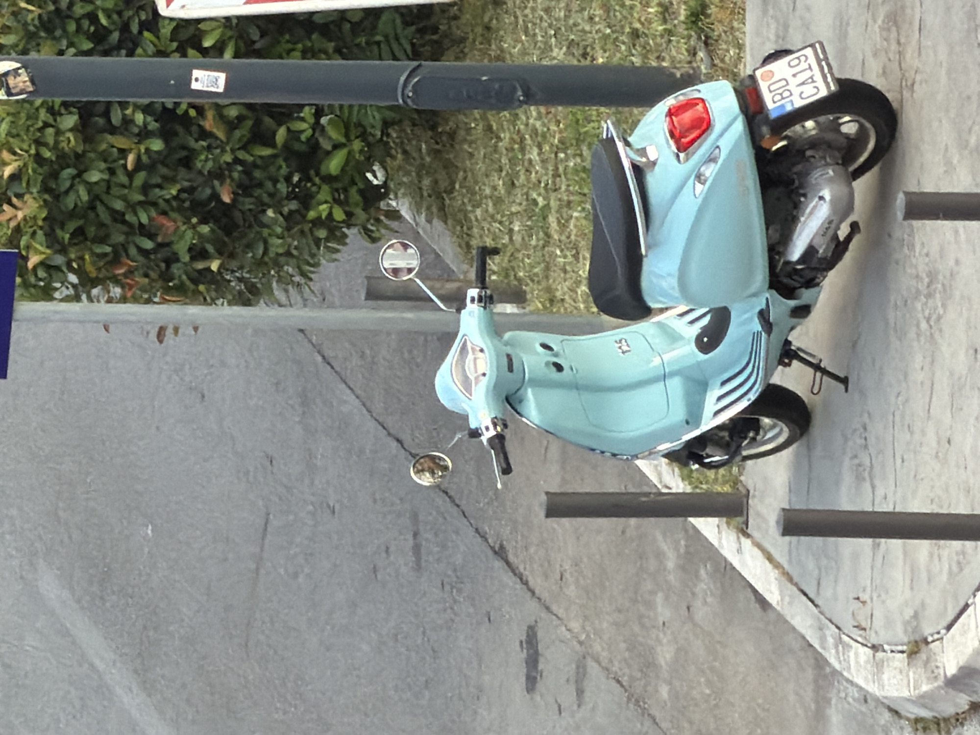 The image shows a light blue Vespa scooter parked on a concrete sidewalk next to a road. The scooter is facing to the right, with its rear to the viewer. The license plate reads "BD CA19". A black pole stands to the right of the scooter, in front of a dense green hedge. The sidewalk is bordered by a curb and a grassy area. In the background, a grey paved surface, likely a street, is visible. The scene appears to be outdoors in an urban environment, potentially Budva, Montenegro, as indicated by the "BD" on the license plate which is a common prefix for Budva vehicle registration plates.