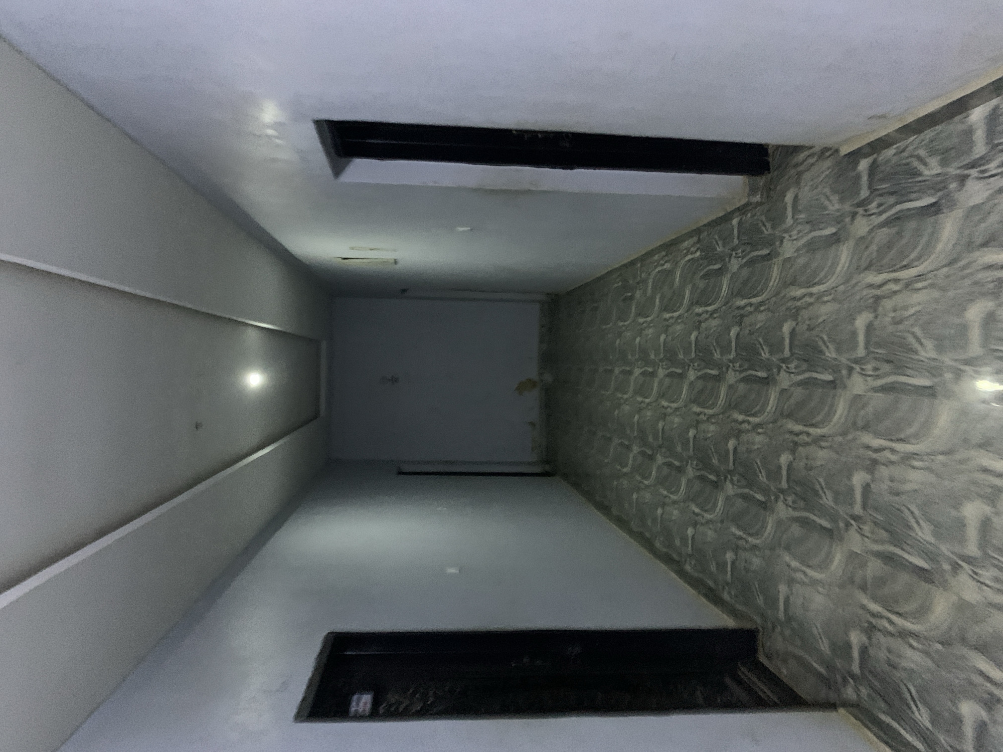 This image depicts a long, dimly lit interior hallway within a building located in Kabong, Nigeria. The hallway features light grey-blue painted walls on both sides. On the left wall, a dark, possibly black, door is visible in the foreground, partially ajar. Further down the hallway, on the right wall, another dark door is closed. The ceiling is white with a recessed central panel extending the length of the corridor, containing at least one illuminated circular light fixture towards the far end. The floor is covered with grey and white patterned tiles, exhibiting a swirled, abstract design. Visible on the lower sections of the right wall towards the distant end of the hallway are patches of discolored or peeling paint. Small, light-colored rectangular fixtures, possibly light switches or outlets, are present on both walls. No individuals, actions, or interactions are visible; the hallway appears empty.