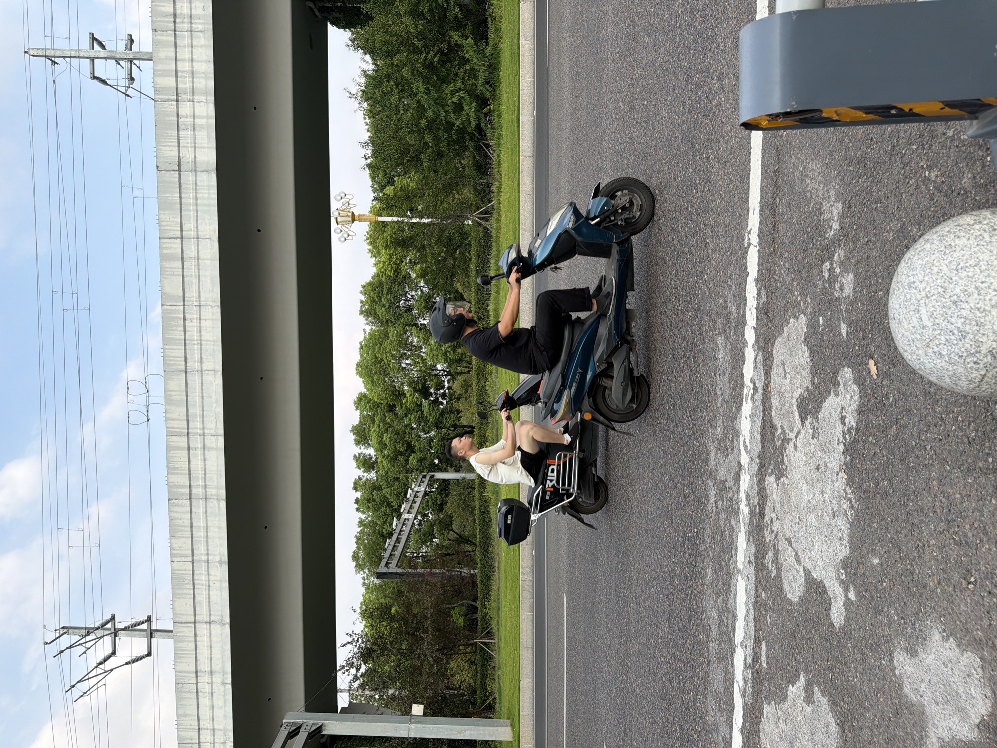 The image shows two individuals on electric scooters riding on an asphalt road. The person in the foreground on the right is wearing a black helmet and black clothing. The person on the scooter in the background to the left is wearing a white sleeveless shirt and black shorts. They are riding under an elevated concrete overpass with power lines and utility poles visible overhead. Trees and green grass line the side of the road. The scene appears to be in an urban or suburban setting in Jiangjin District, Chongqing, China.