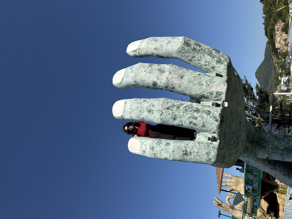 A female subject stands within the palm of a monumental hand sculpture, located at VinWonders Nha Trang on Hon Tre Island, Nha Trang, Vietnam. The subject has dark, shoulder-length hair, wears dark sunglasses, a red short-sleeved top, and dark trousers, facing directly towards the observer. The colossal hand, appearing to be crafted from a textured, teal-green material with contrasting white fingertips, occupies the central mid-ground. The upper portion of the image is dominated by a clear, unclouded blue sky. In the lower-left background, a secondary architectural feature resembling another large hand sculpture, integrated with a walkway and a small sheltered structure, is visible, with a distant figure standing near it. The broader background reveals rolling green hills, scattered white buildings, and areas of exposed earth. The scene is brightly illuminated by direct sunlight.
