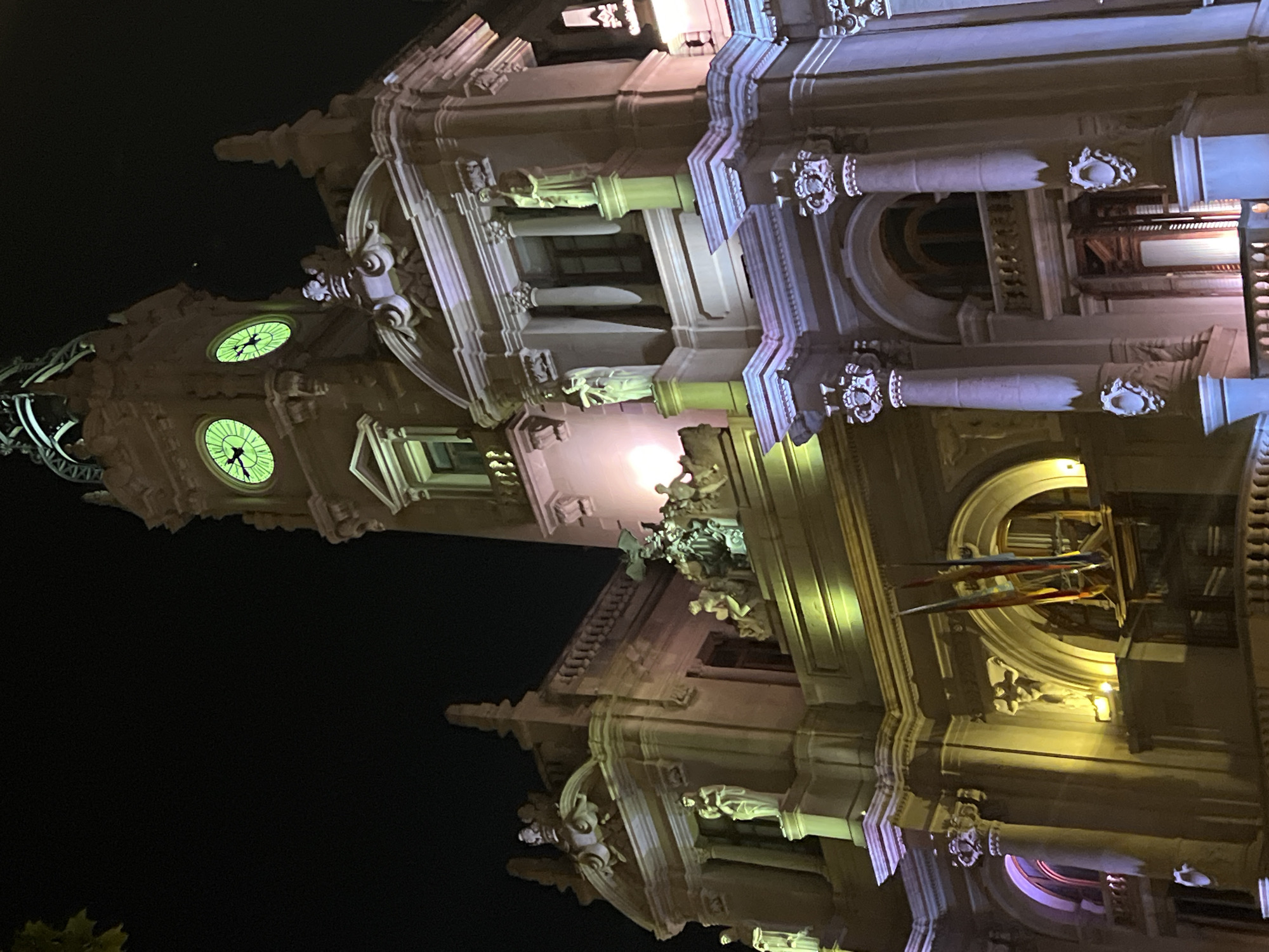 The image shows a building at night in Valencia, Spain. The building is illuminated and has a clock tower with two visible clock faces. The building is highly ornamented with statues and architectural details. A set of three flags is visible, hanging beneath an archway. The sky is black, suggesting it is late at night.