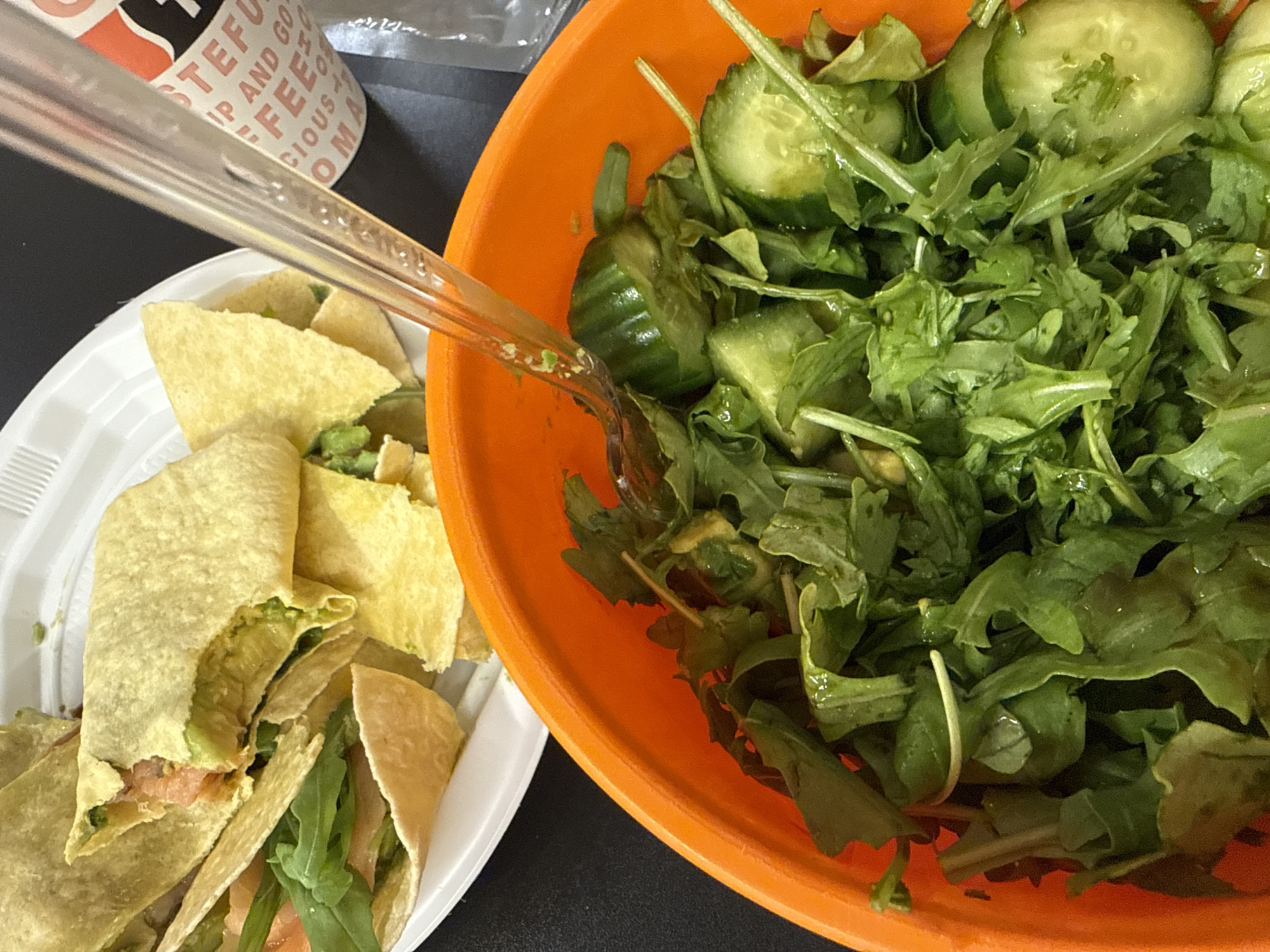 The image captures a casual meal set on a dark surface, featuring two main dishes. In the foreground and right side, a vibrant orange bowl holds a fresh green salad, primarily consisting of arugula (rocket leaves) and sliced cucumbers, lightly coated with what appears to be dressing. A clear plastic fork or spork, with remnants of food on it, rests across the rim of the orange bowl and extends towards a white disposable plate on the left.
The white plate contains several small, soft wraps or tacos, some appearing to be filled with mashed avocado or guacamole. Another visible wrap reveals layers of what looks like reddish-pink smoked salmon or deli meat alongside green leaves, possibly more arugula. Scattered around the wraps are pieces resembling tortilla chips or broken wrap pieces.
In the blurred background, a white paper cup is partially visible, featuring text that reads "STEFUL", "SIP AND GO", "FFEE", and "CIOUS AROMA", suggesting it's a coffee cup. The overall scene depicts a casual dining experience, possibly lunch, given the fresh ingredients and the type of meal. The use of disposable plates and plastic utensils indicates a non-formal setting, potentially a takeaway, office meal, or picnic. No people are visible. The location context provided is Tel-Aviv, Israel, though no visual cues in the image confirm this.