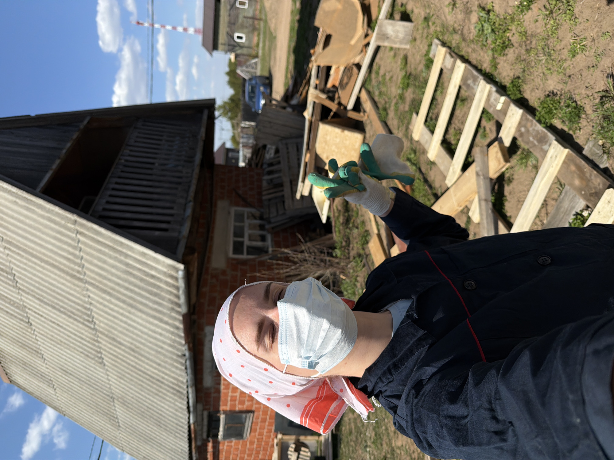 A woman wearing a light blue surgical mask, a white headscarf with red polka dots, a navy blue work jacket, and white gloves with green fingertips stands outdoors. She is positioned near some newly constructed wooden frames. Behind her is a building with a corrugated metal roof and a red brick facade. Discarded wood and other debris are scattered on the ground. Further in the background, other buildings and a red and white striped tower are visible. Based on these details, the location is identified as the city of Chaikovsky, Russia.