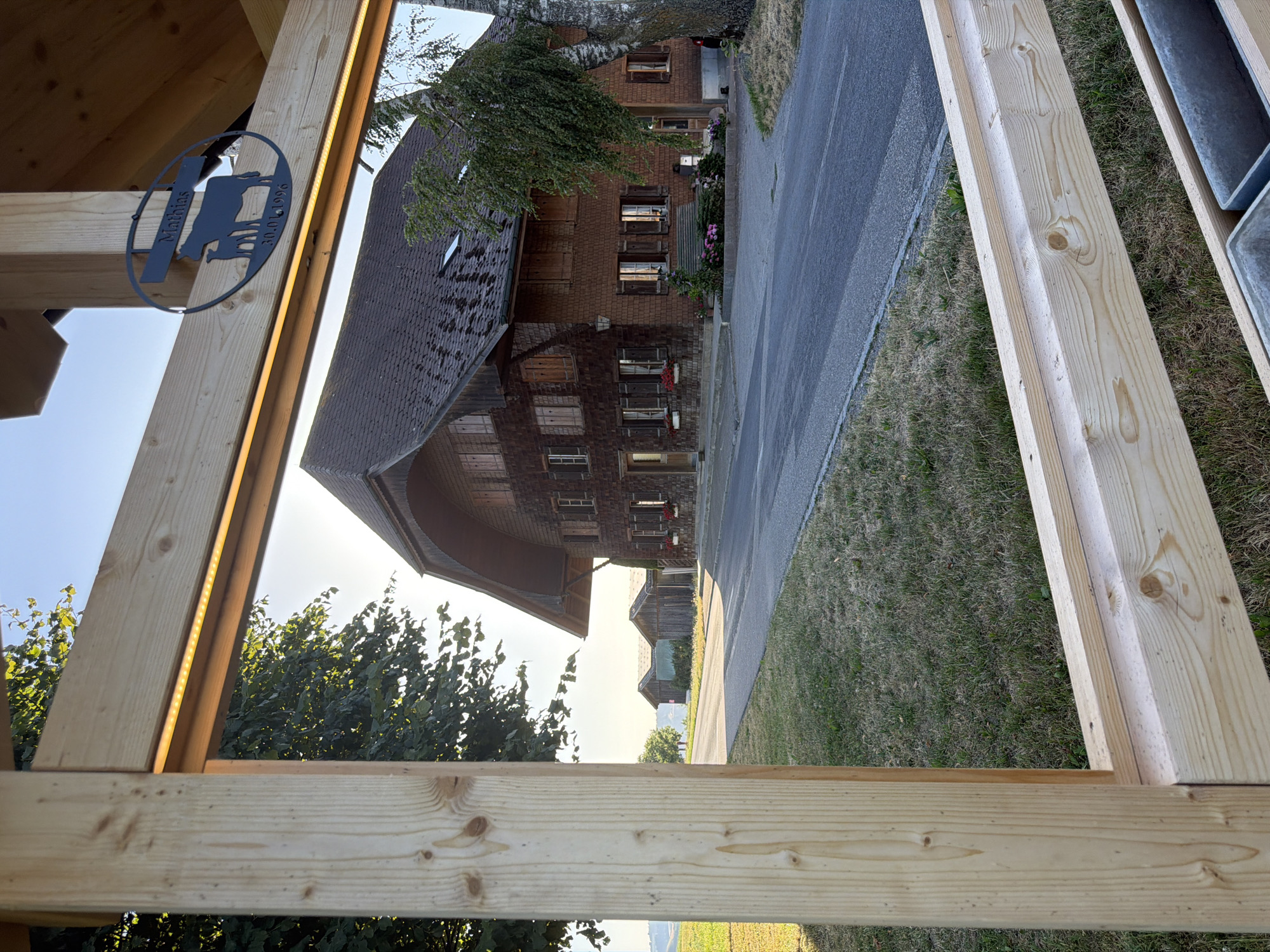 A wooden frame, partially constructed, offers a view of a traditional Swiss farmhouse with a steep, shingled roof and dark wooden shutters on its windows. The house is surrounded by trees and a paved road. In the foreground, the unfinished wooden structure includes two vertical posts and a horizontal beam with a metal silhouette attached. This silhouette depicts a cow and calf, with the name "Mathias" and the date "30.01.1996" inscribed below. The scene is bathed in the warm light of a setting sun, casting long shadows and illuminating the sky with a soft glow. The location is identified as Ufhusen, Switzerland.
