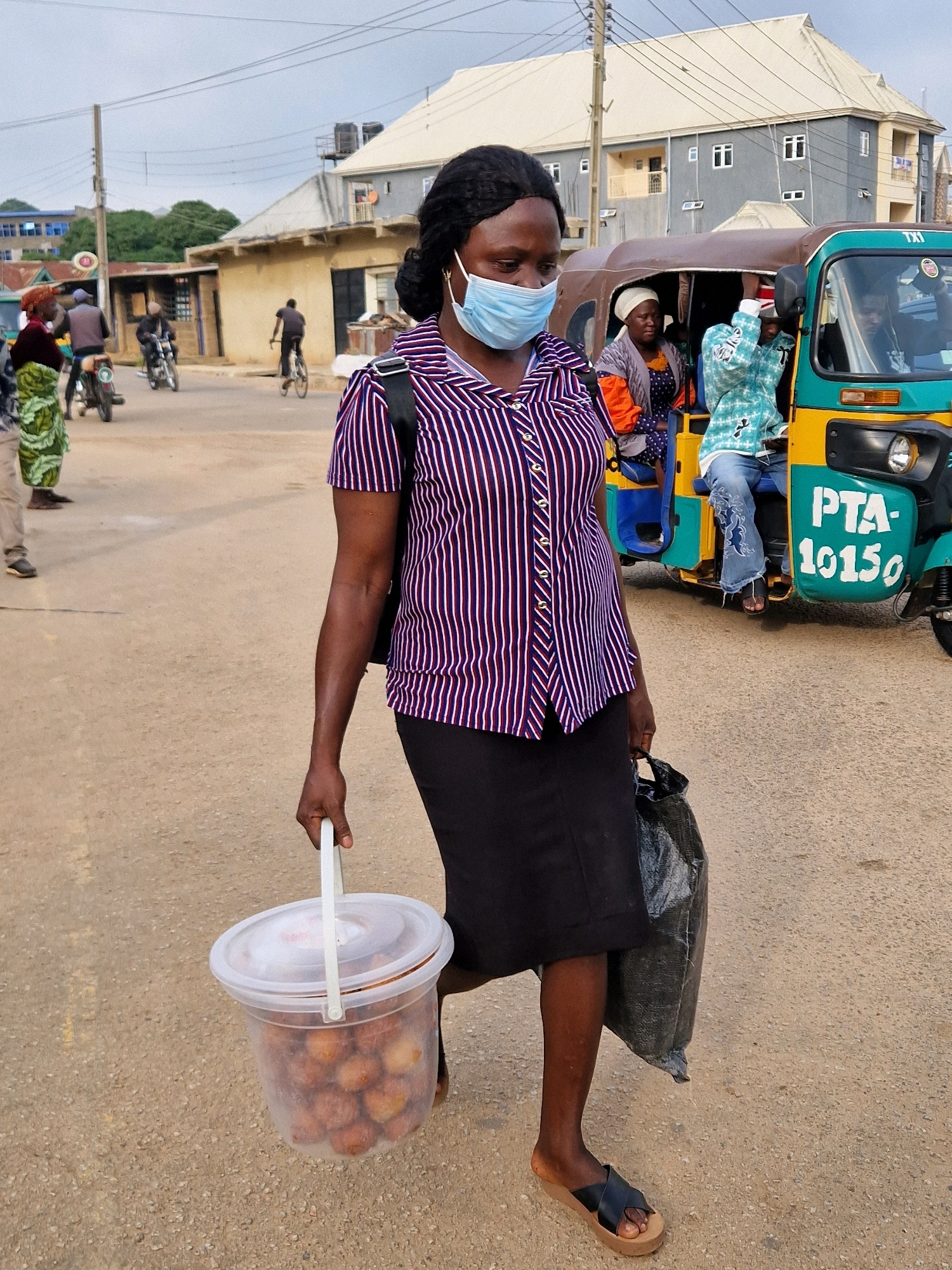 In New Stadium, Nigeria, a woman wearing a striped blouse and a face mask walks along a dusty road. She is carrying a clear plastic bucket filled with food items in her left hand and a black plastic bag in her right. Behind her, a trishaw vehicle, identified with "PTA-10150" painted on its side, is stopped. The environment appears to be a semi-urban setting with buildings and overhead wires visible in the background. Several other people are present, some on motorcycles and one on a bicycle, indicating a bustling thoroughfare.