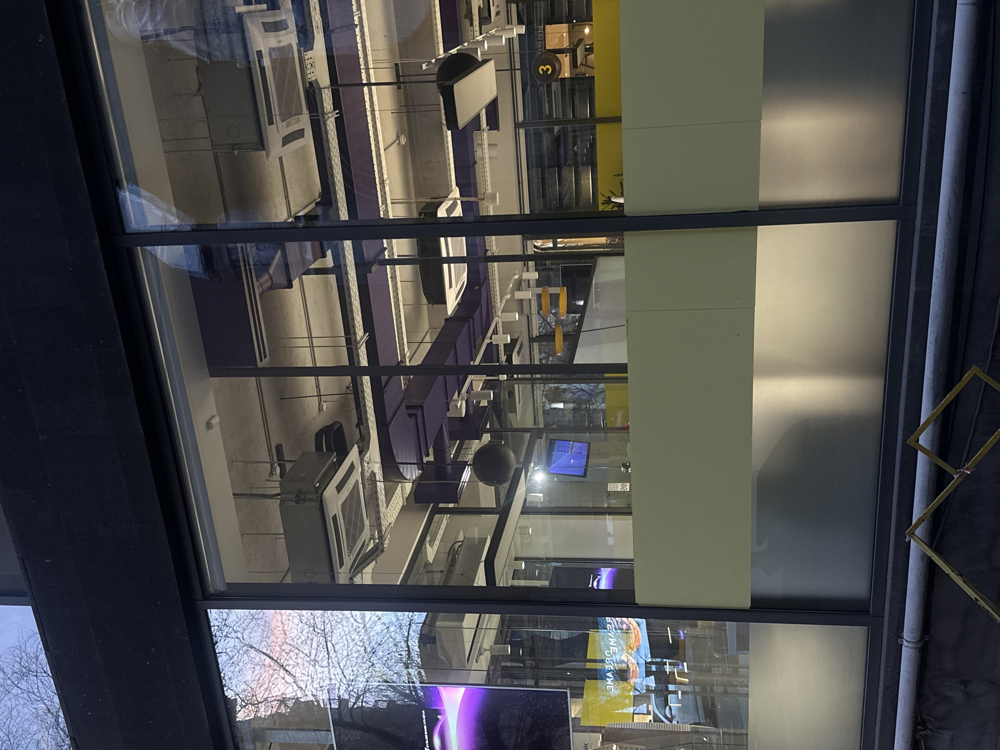 The image depicts an interior reflected in a window, likely in Yerevan, Armenia. The interior appears to be a modern office space, with suspended air conditioning units, overhead lighting, purple structural supports, and office furniture. A spherical object hangs suspended from the ceiling. A television screen can be seen. The window reflects trees and a building. Outside the window is a building exterior. The window itself consists of multiple panes with dark frames.