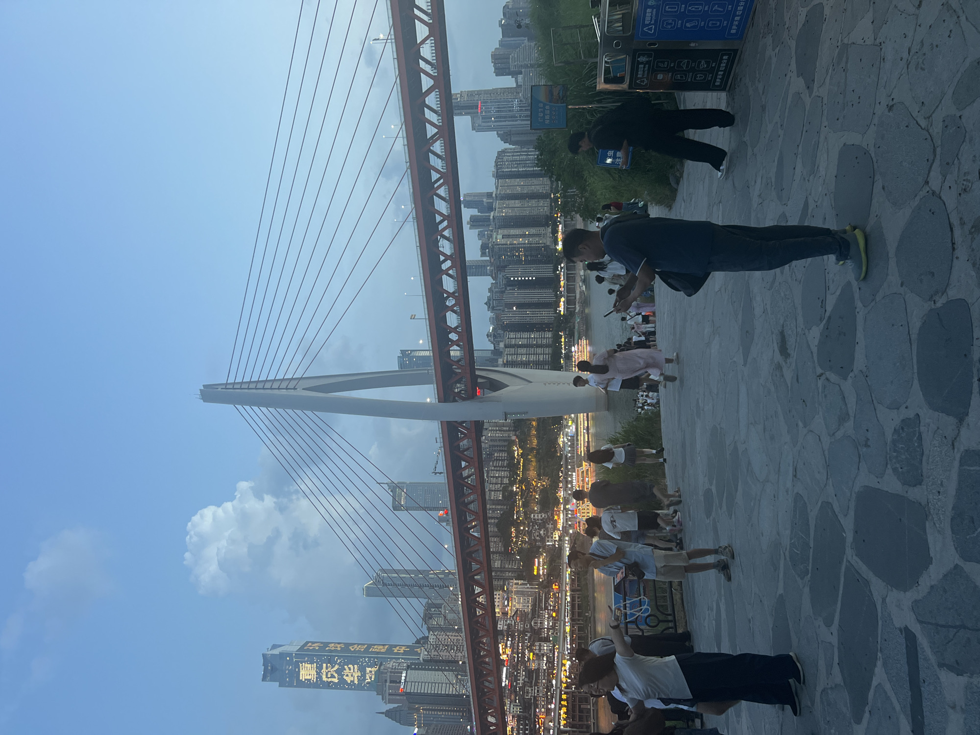 This is a medium-shot, eye-level photograph of a pedestrian walkway in Jiangbei Cheng Street, China, likely taken in the early evening. The walkway is paved with large, irregular stone slabs. In the foreground, several people are walking and standing. A man in a dark blue t-shirt and dark pants is looking at his phone. To his left, a woman in a white t-shirt and dark pants is facing away from the camera with her arms outstretched. Further in the background, more people are scattered across the walkway and near a body of water.
Dominating the mid-ground and background is a large cable-stayed bridge with a prominent white pylon and a red superstructure. Beyond the bridge, a densely built urban skyline rises, characterized by numerous skyscrapers and buildings illuminated by artificial lights. Some of these buildings have illuminated text on their facades. A river flows in front of the cityscape, with boats visible on its surface. The sky is a dusky blue, with scattered clouds. Two trash receptacles are visible on the right side of the frame, with Chinese text indicating recycling information.