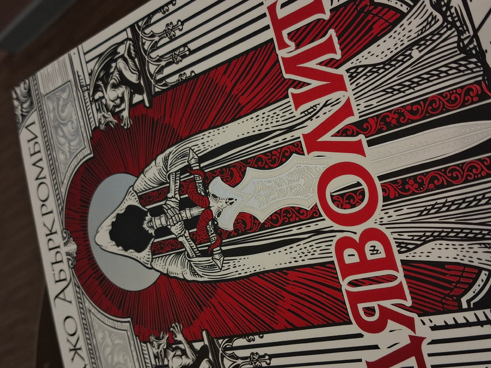 The image depicts a book cover with a stylized illustration. The cover features a large, central design of a skeletal figure draped in fabric, holding a sword, with a silver circular shape behind the skull. The background has red, black, and white lines radiating outward from the figure, and a decorative border with gargoyle-like figures. Red Cyrillic text is superimposed over the illustration. The text "ЖО АБЭРКРОМБИ" is printed along the left edge of the book cover. The location is unspecified.