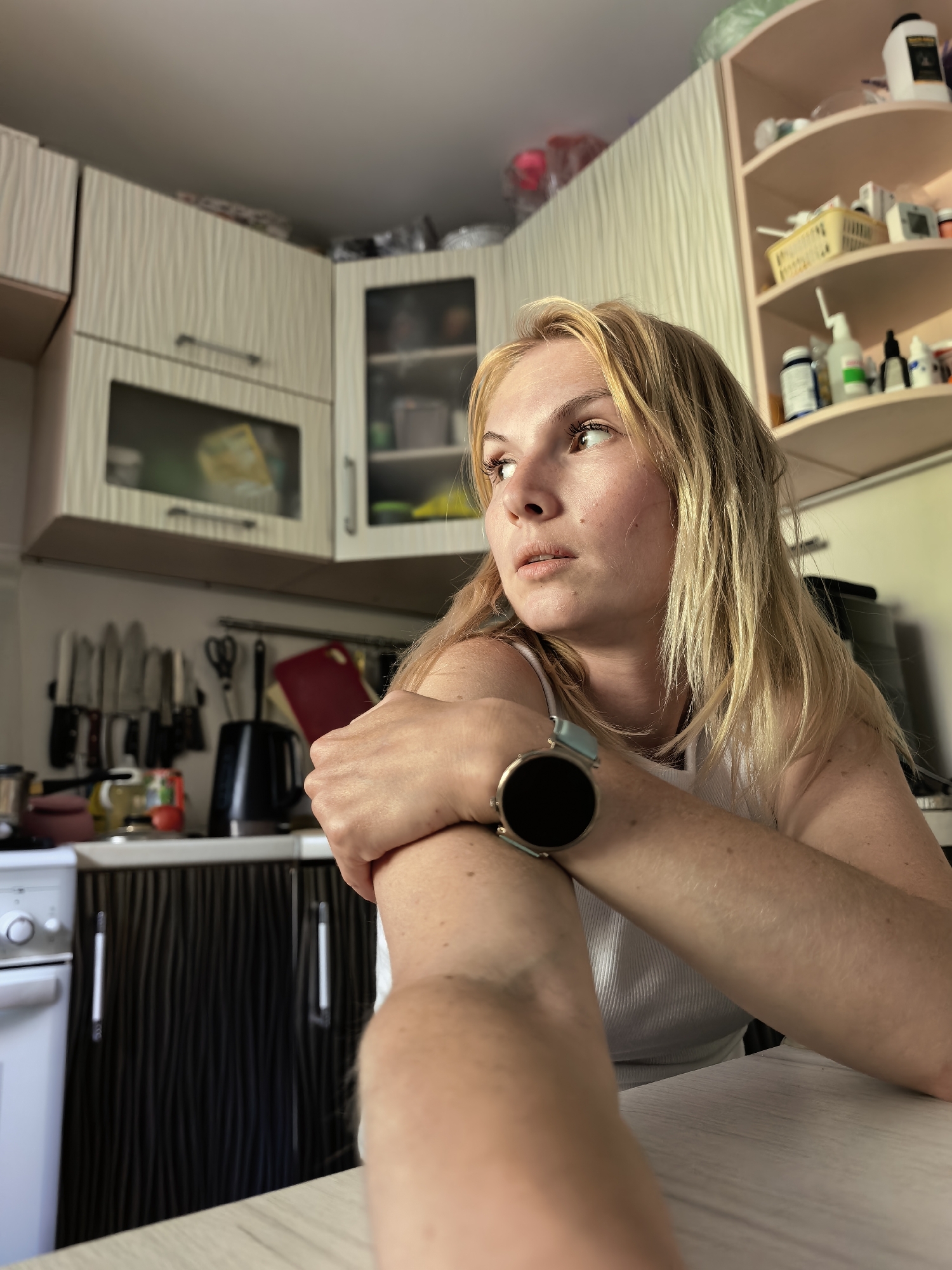 A young woman with blonde hair is sitting at a table in a kitchen. She is wearing a white tank top and a smartwatch on her left wrist. Her arms are crossed in front of her chest as she looks away from the camera. The kitchen has light-colored cabinets and a dark-colored countertop. There is a stove to the left of the frame, and shelves filled with various items are visible in the background. The lighting suggests it is daytime. The location is identified as Sterlitamak, Russia.