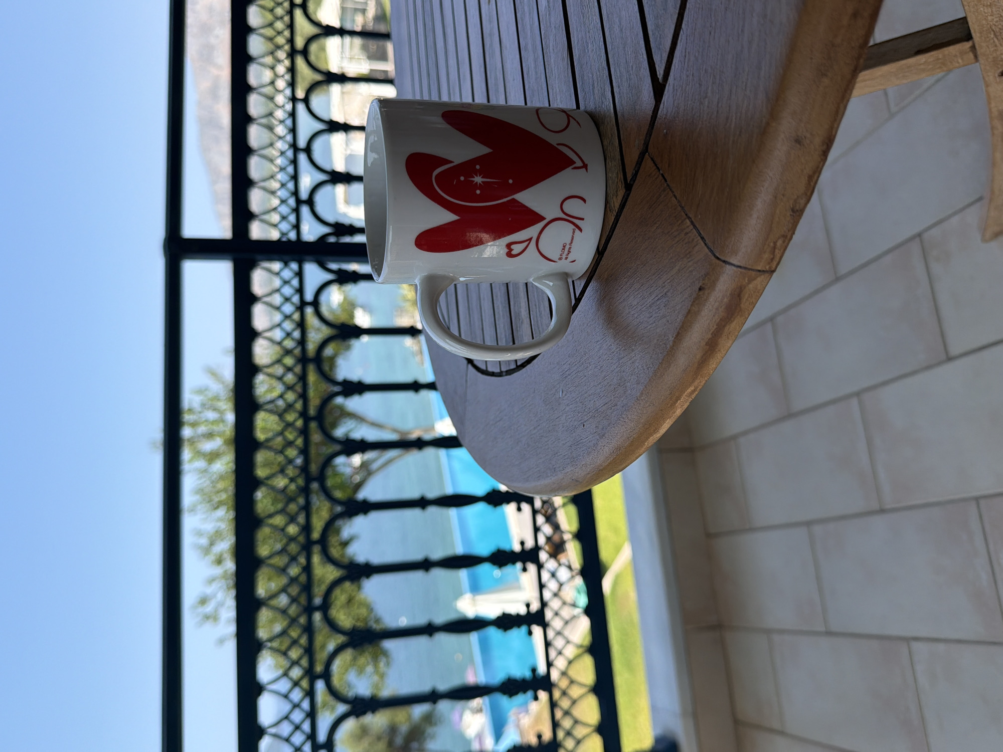 A white mug with red heart designs sits on a wooden table on a balcony overlooking the sea. The mug has two large red hearts and smaller red heart designs. The balcony has a black metal railing with an ornate pattern. Beyond the railing, a bright blue swimming pool is visible, with lounge chairs and umbrellas. In the distance, the clear blue sea stretches out to meet a hazy, mountainous coastline under a clear blue sky. The tiled floor of the balcony is visible in the foreground. The overall scene suggests a relaxed, vacation setting in a coastal area.