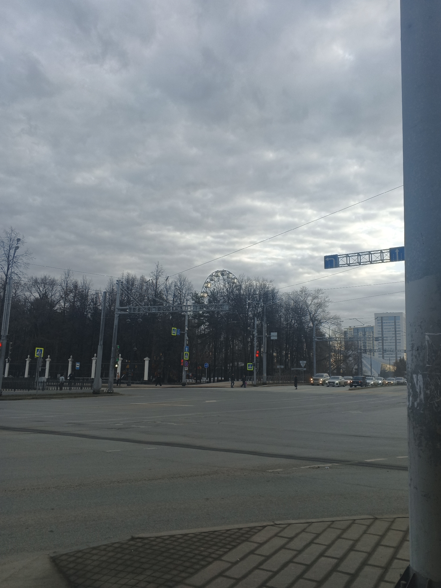 The image shows a street scene in Perm, Russia, on an overcast day. In the foreground, a wide intersection is visible, featuring multiple lanes and crosswalks, with light vehicular traffic. The road is dry and appears well-maintained. In the background, a park lines the road, with bare trees and a decorative fence that runs along its border. A Ferris wheel is partially visible behind the park's trees. Several buildings can be seen further in the distance, including one high-rise structure. Traffic lights and road signs are positioned on either side of the road. A utility pole is visible in the right foreground. Pedestrians are present, crossing the street.