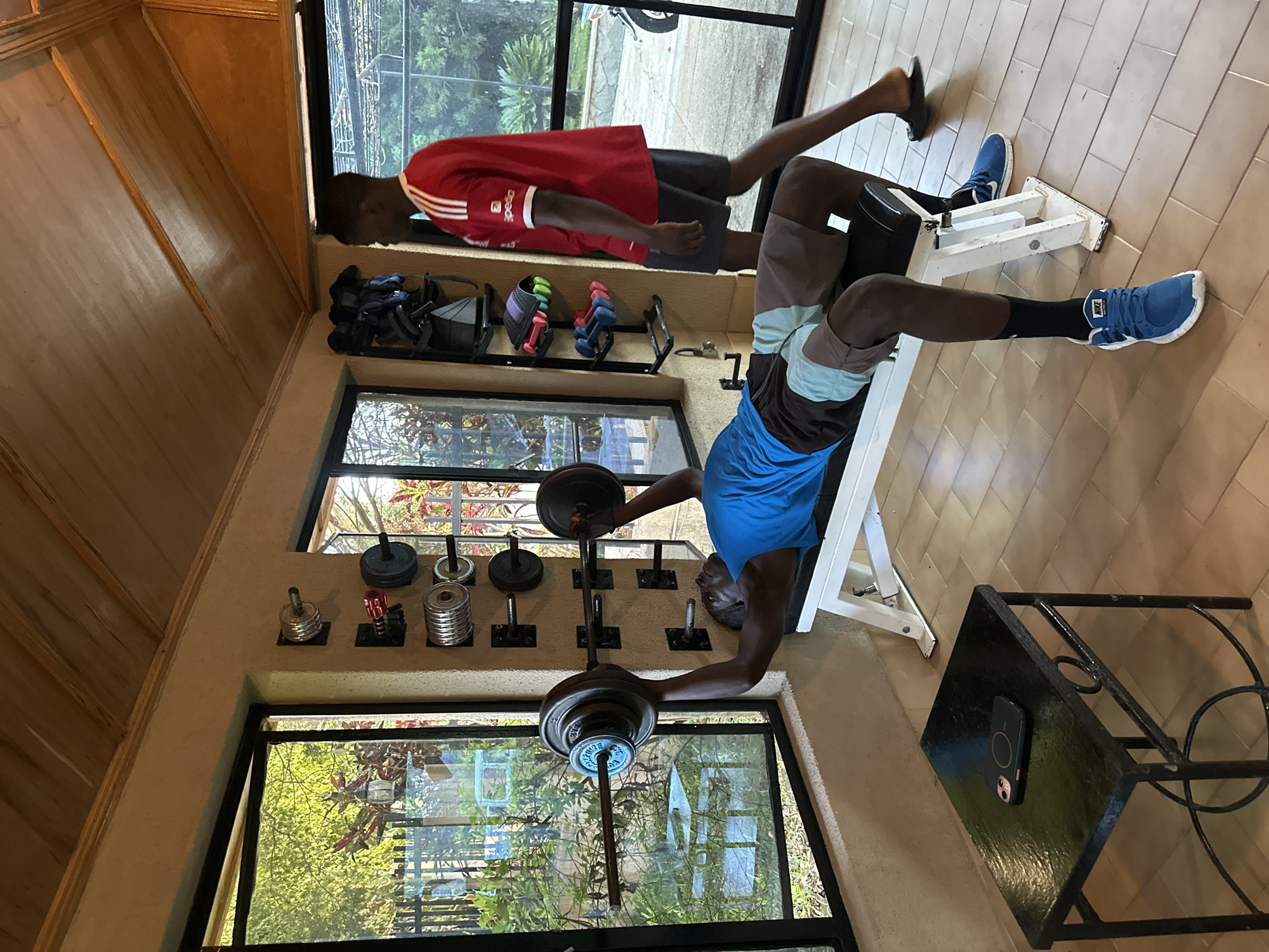 A gym setting in Jos, Nigeria is seen in the image. A man wearing a blue t-shirt and shorts is lifting a barbell on a bench. Another man in a red jersey is standing in the background, looking out the window. Dumbbells, weight plates, and a phone on a table are also visible. The room has large windows showing green foliage outside. The ceiling is wooden.