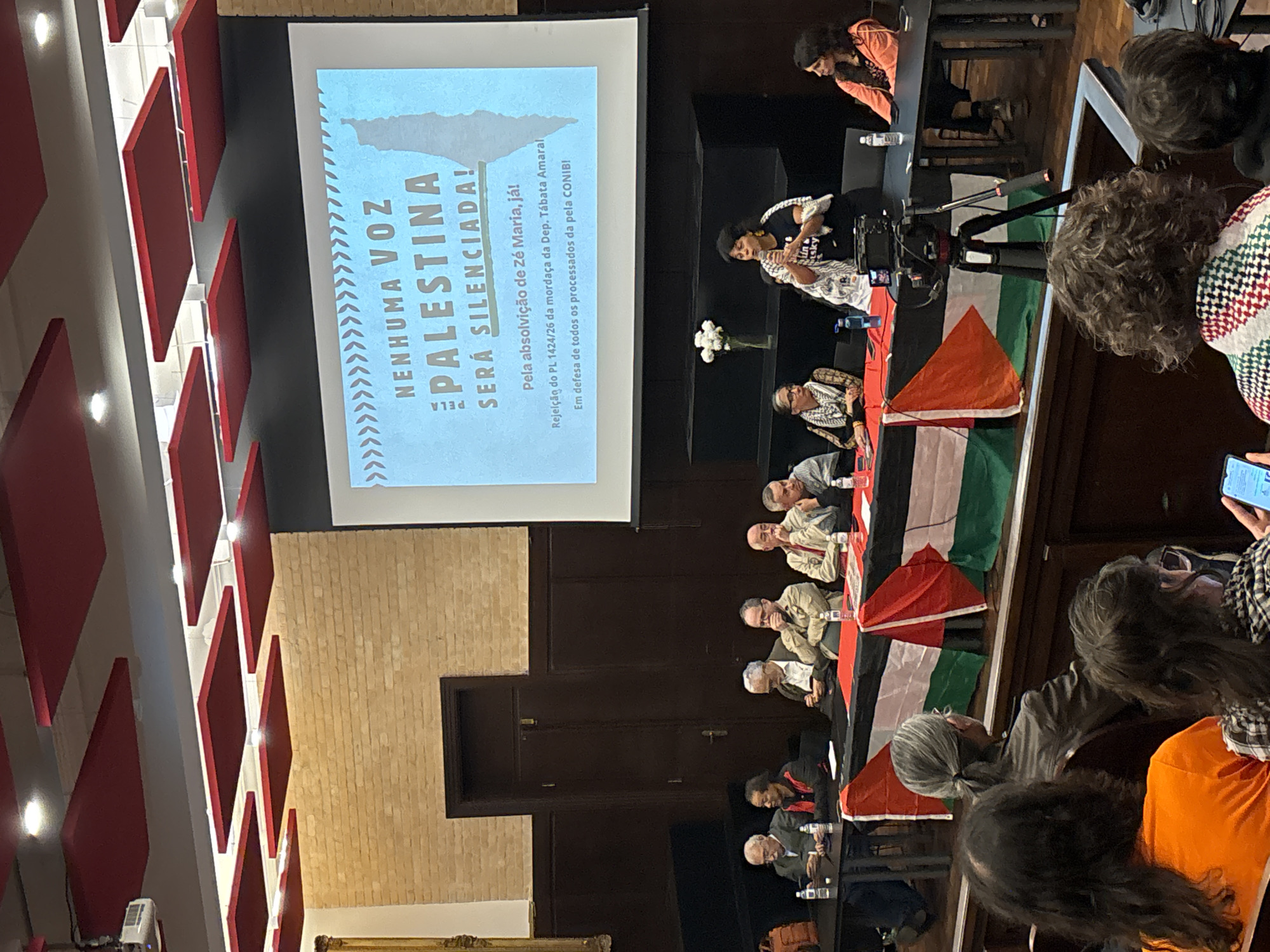 The image captures a room in São Paulo, Brazil, where a presentation with a pro-Palestinian message is being held. The text on the screen states, "Nenhuma voz da Palestina será silenciada!" which translates to "No Palestinian voice will be silenced!". It also mentions support for Zé Maria and opposition to PL 1424/26 by Deputy Tábata Amaral. A panel of individuals sits at a table draped with Palestinian flags. People are sitting in the audience, some taking pictures with their phones. A woman stands by the table, and another is positioned behind her in what appears to be a wheelchair. A camera is set up, potentially recording the event.