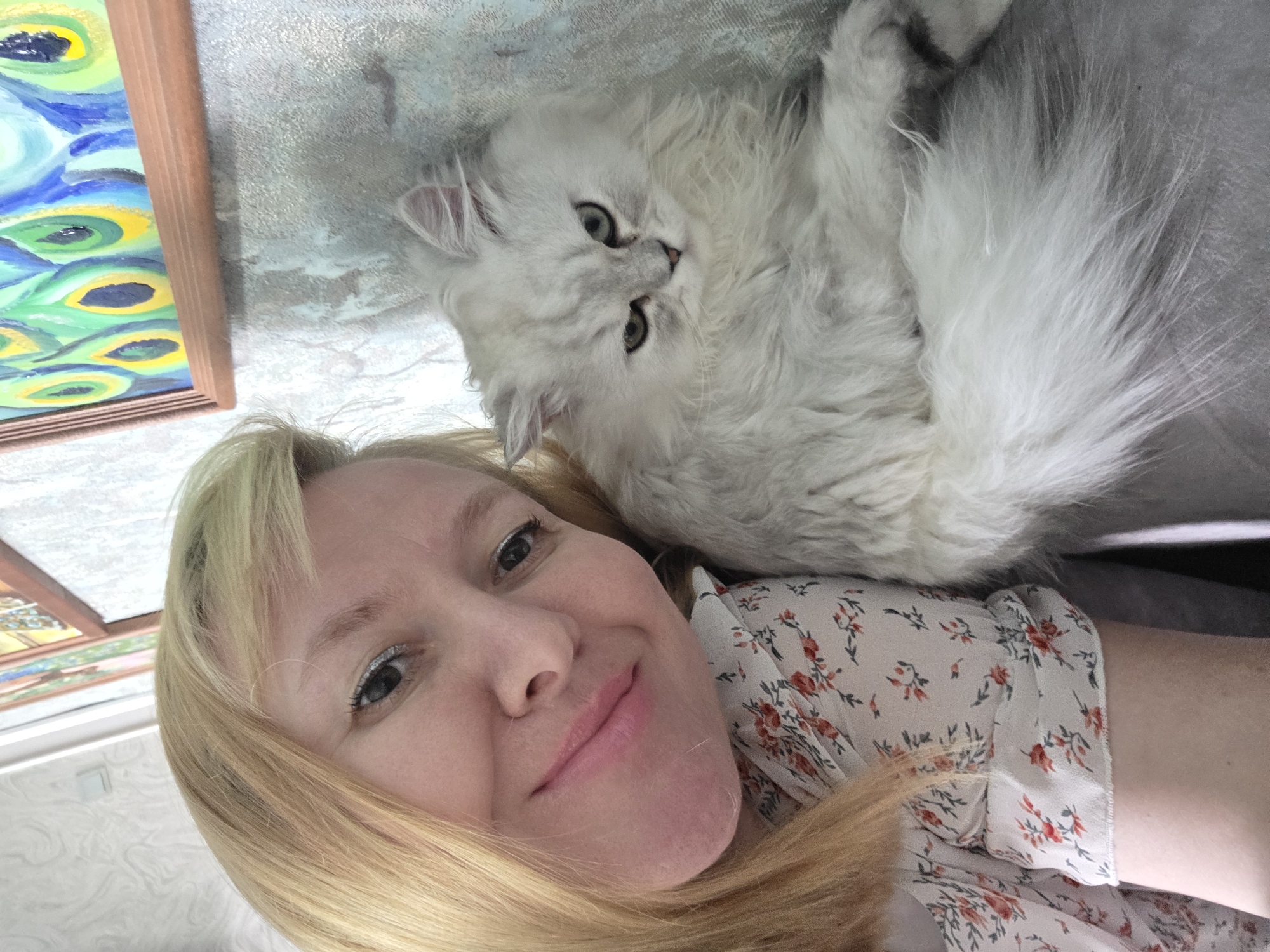 The image shows a selfie of a blonde woman with a white cat. The woman is smiling and the cat is looking at the camera with a slightly grumpy expression. They are both sitting on a gray surface. In the background, there is a painting of peacock feathers. The scene appears to be indoors, in a home environment. The location is the city of Novocheboksarsk, Russia.