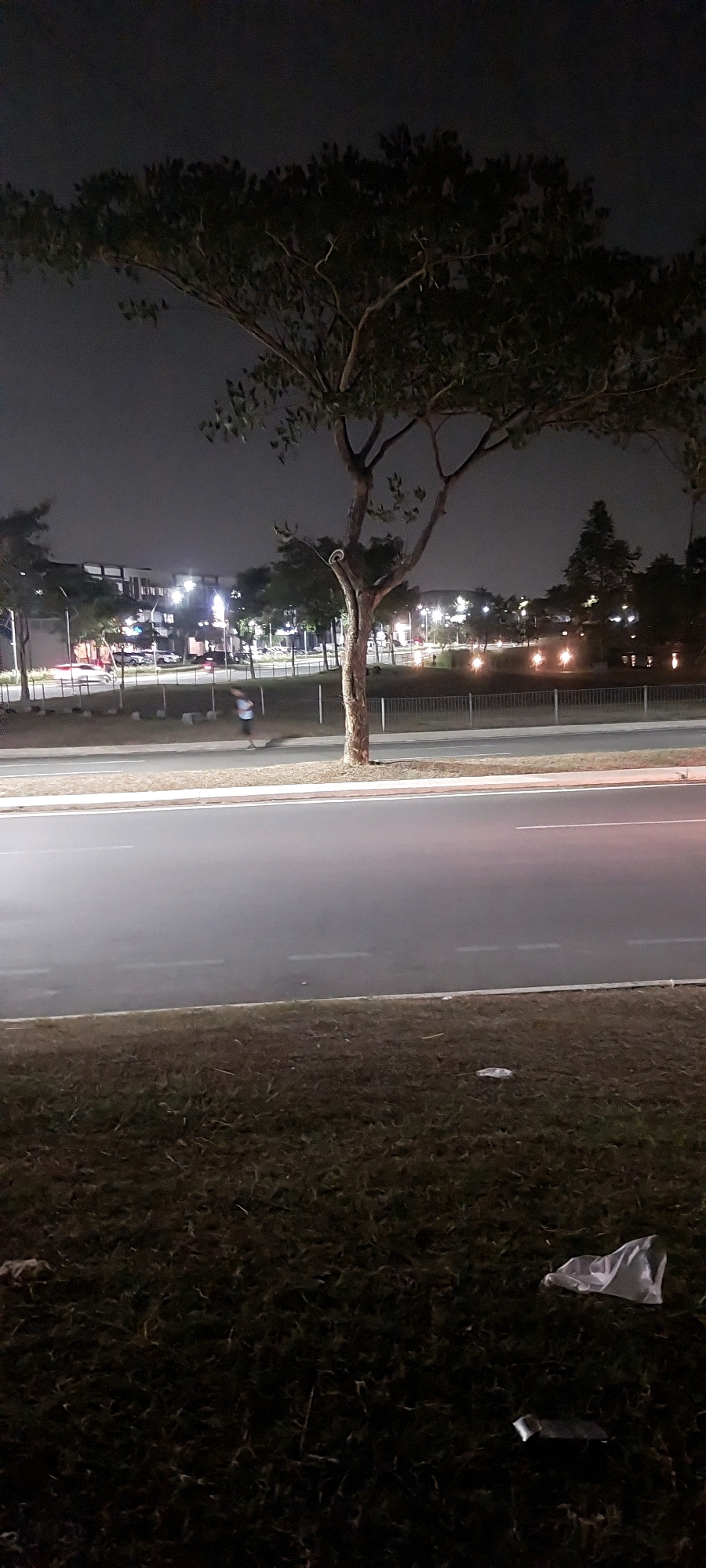 The image depicts a night scene along a road in Medang, Indonesia. A lone person, appearing as a blurred figure, is visible walking along the side of the road. The road itself is wet and reflects the ambient light from streetlights and buildings in the background. Across the road, there are trees, a fence, and low-lying vegetation. In the distance, modern buildings with illuminated windows and streetlights are present, suggesting an urban or semi-urban environment. The foreground consists of a grassy area with some litter, including a crumpled white plastic bag and a metallic object. The sky is dark and overcast, typical of nighttime conditions.