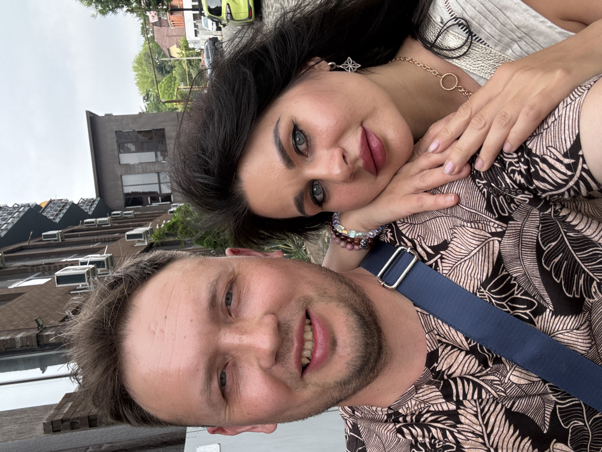 A man and a woman are captured in a selfie outdoors, with a building and trees visible in the background. The man, with short brown hair and a beard, is smiling broadly, showing his teeth. He is wearing a patterned t-shirt and a blue strap across his chest. The woman, with long dark hair, has her head resting on the man's shoulder and is looking towards the camera with a neutral expression. She is wearing makeup and jewelry, including earrings and a necklace. The background suggests an urban setting, possibly a street or a courtyard in Sochi, Russia, given the architectural style and the presence of a car on the street. The sky is overcast, indicating a cloudy day.