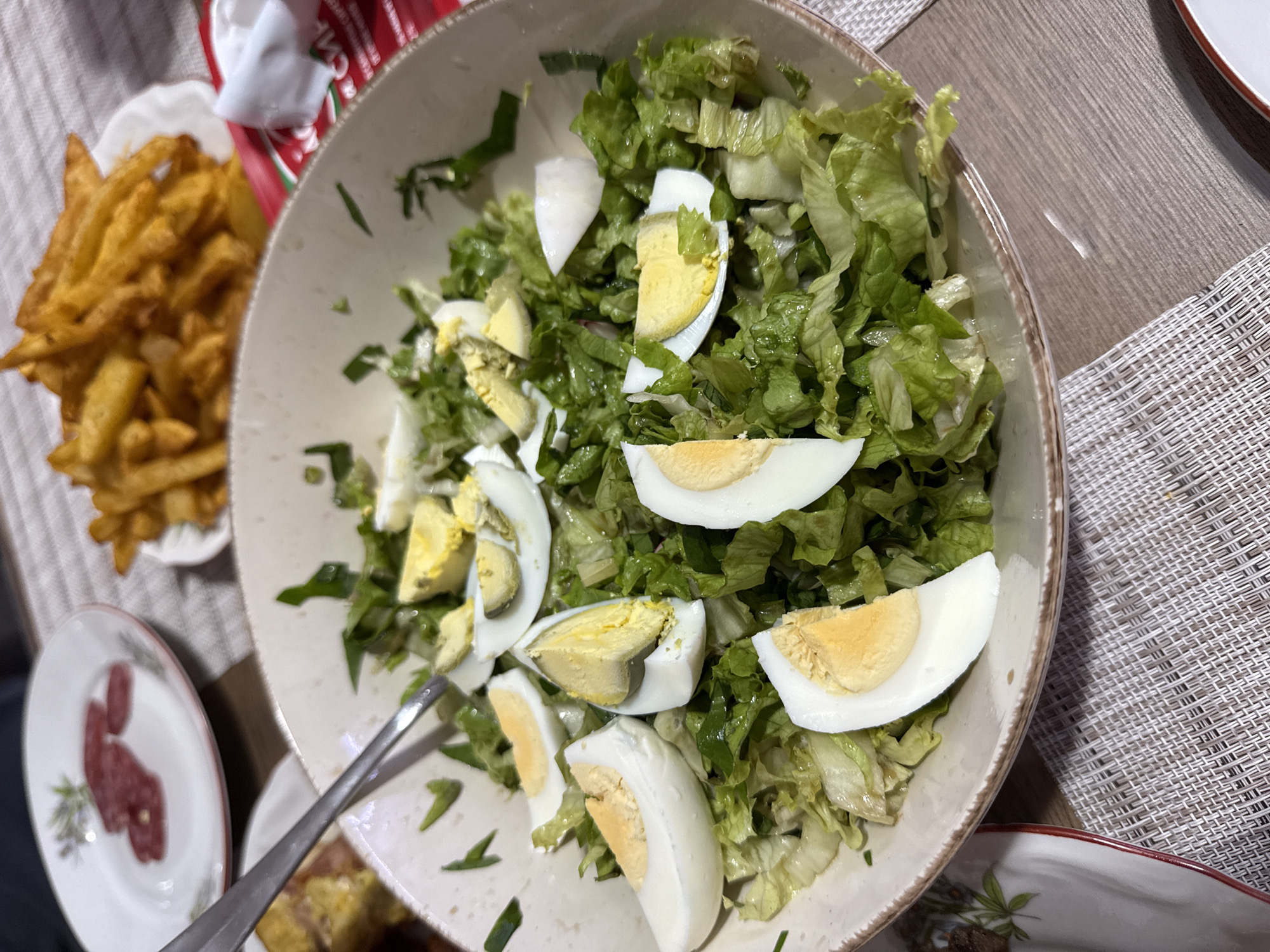 The image shows a table setting with a bowl of salad containing lettuce and sliced hard-boiled eggs as the main focus. To the left of the salad bowl, there is a small plate of french fries and another small plate with what appears to be slices of meat. A serving spoon is visible within the salad bowl. The setting appears to be indoors, likely in a home or restaurant. The table has a textured placemat. The location is Sofia, Bulgaria.