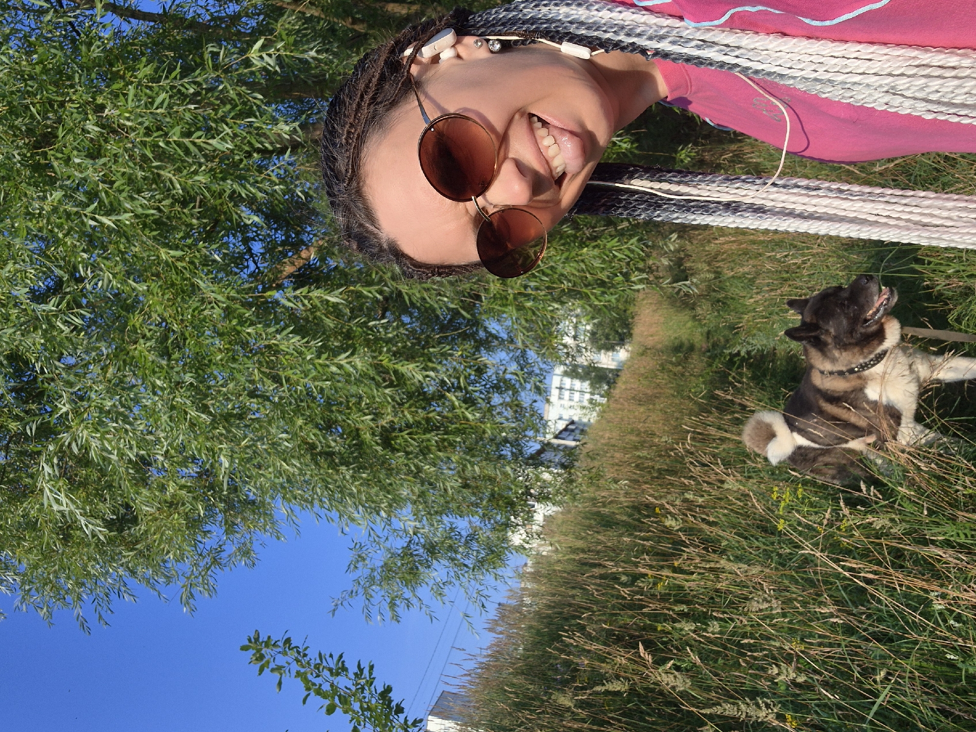 The image shows a woman with braided hair wearing sunglasses and a pink shirt, sticking her tongue out. She is wearing white earbuds. Beside her, a dog with brown and white fur is walking on a leash. They are in a grassy area with trees and buildings in the background. The woman appears to be taking a selfie. The scene is outdoors, under a clear blue sky. This image was taken in Chaikovsky, Russia.
