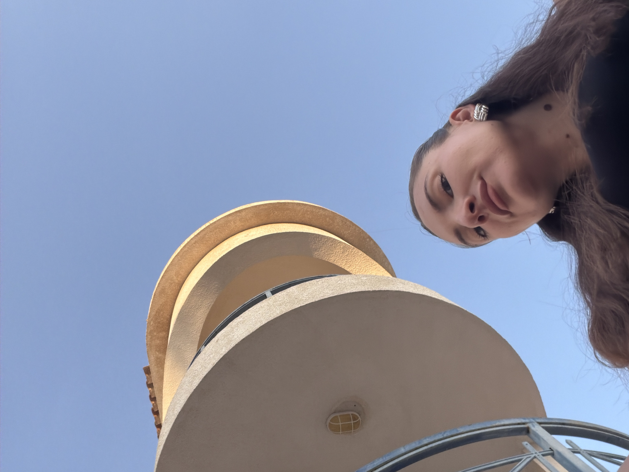A low-angle outdoor shot captures a young woman looking up at the camera with a building structure and clear blue sky in the background. The woman, positioned in the lower right of the frame, has long brown hair and is wearing large silver earrings and a black top. The building, a pale yellow cylindrical structure with curved balconies, is seen from below, with its top section featuring terracotta roof tiles. A small oval light fixture is visible on the lower part of the building. The composition suggests a selfie taken outdoors, possibly in a resort area given the architectural style, which is consistent with some of the modern structures found in Sharm El Sheikh, Egypt, a popular tourist destination known for its coastal resorts and clear skies.