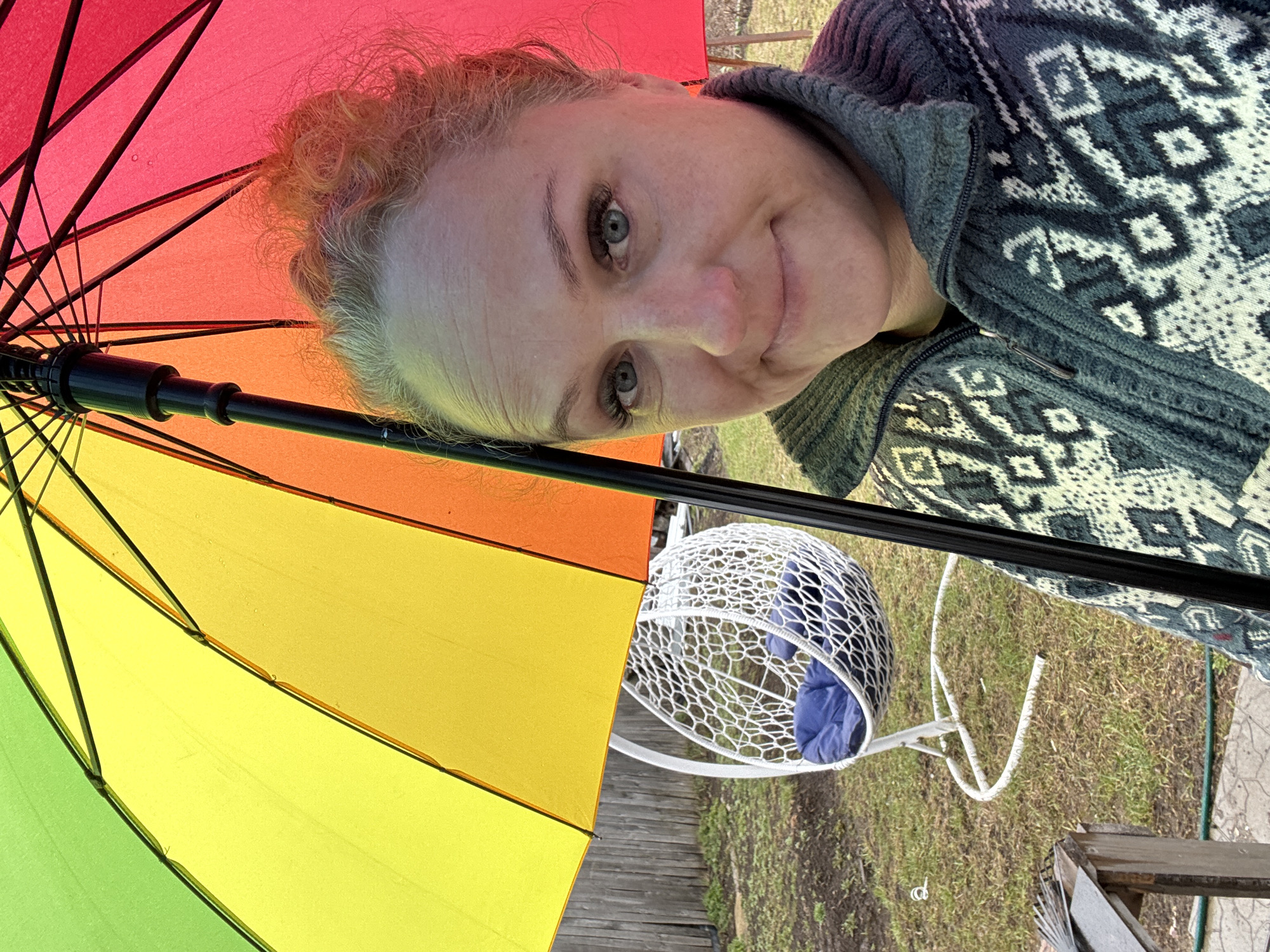 A woman is under a rainbow umbrella, looking at the camera. She has light skin, blue eyes, and light blonde hair. She is wearing a gray and white patterned sweater. Behind her is a white egg chair with blue cushions, sitting on a grassy lawn. The location is Chaikovsky, Russia.