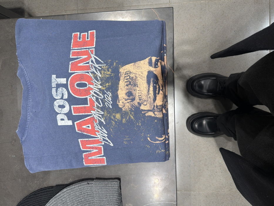 A navy blue t-shirt with "POST MALONE LIVE IN CONCERT 2026" printed in red and white sits on a gray surface. The shirt features an image of Post Malone's face. Below the shirt are two knit hats, one black and one gray. A person wearing black pants and black loafers stands next to the surface, with the tips of their black coat visible. The surface appears to be a table or display in a retail setting.