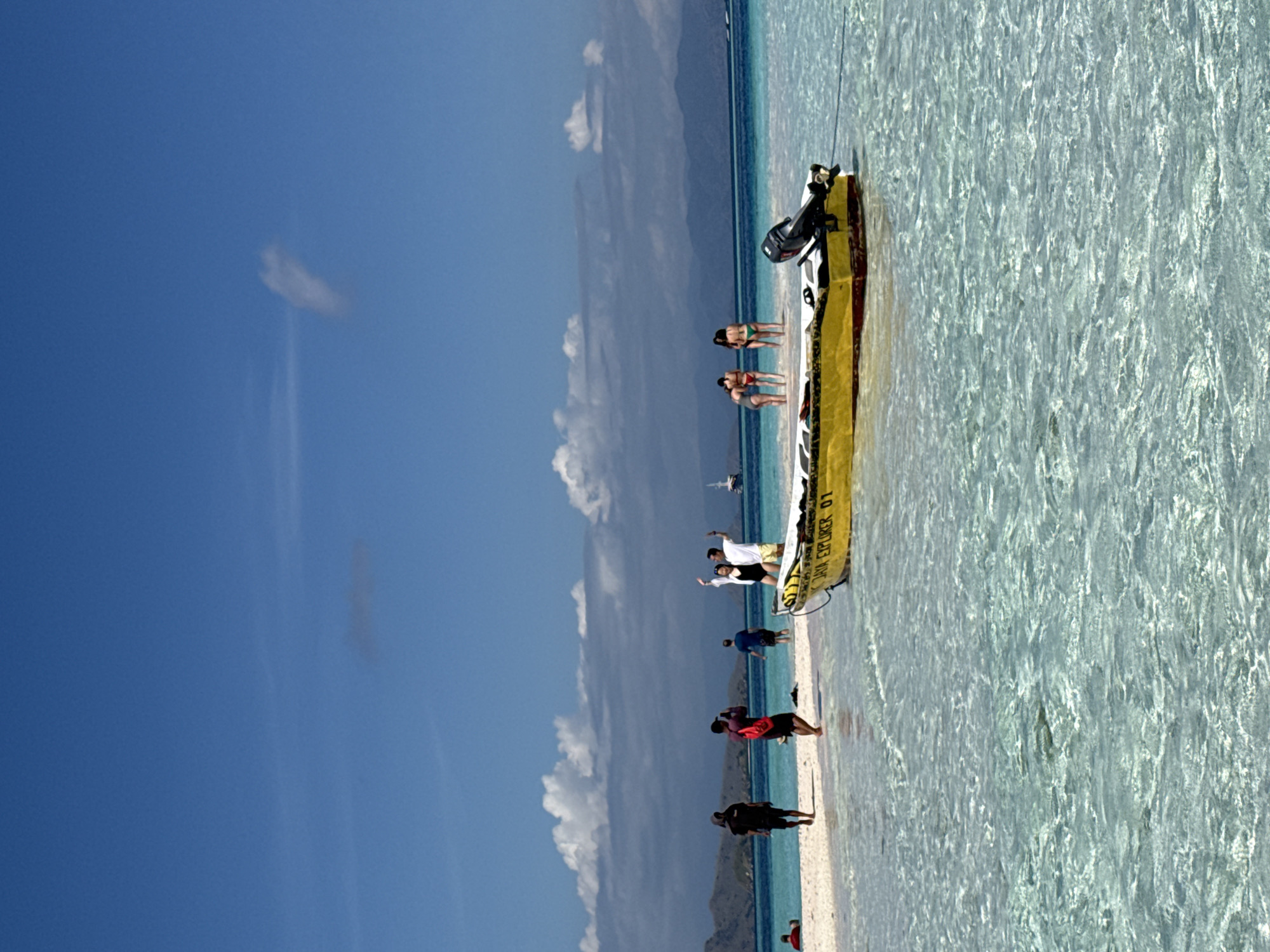 The image depicts a beach scene in Komodo, Indonesia, featuring a long, narrow yellow and white boat beached on a sandbar. Several individuals are positioned along the sandbar, interacting and enjoying the location. The water is clear and turquoise, contrasting with the blue sky and scattered clouds. Mountains are visible in the distance.