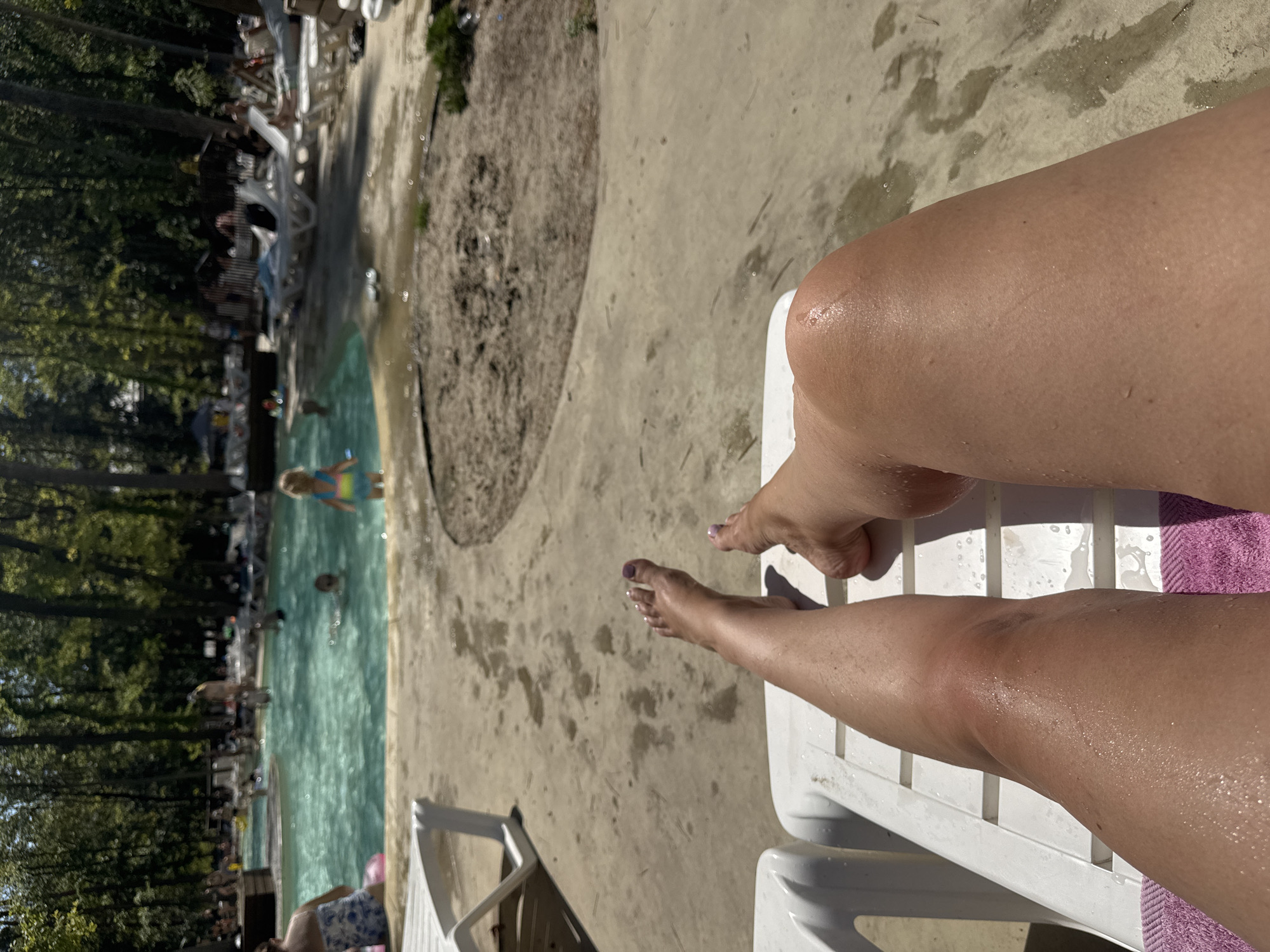 The image shows a first-person perspective of a person relaxing on a white lounge chair with a pink towel underneath. The person's tanned legs are visible, glistening with water droplets. In the background, a busy outdoor swimming pool area is depicted, with trees lining the perimeter. Several people are in and around the pool, including children playing in the water. Lounge chairs are scattered around the pool area, some occupied by sunbathers. The ground around the pool is paved and wet in places, suggesting recent activity. The overall scene depicts a sunny day at a recreational water facility. The provided text indicates the location is Orlivshchyna, Ukraine, but this information is not visually verifiable within the image itself.